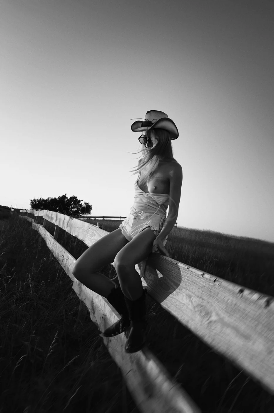 A woman wearing cowboy hat, sunglasses, and boots sitting on a wooden fence in an open field during sunset, black and white photo.