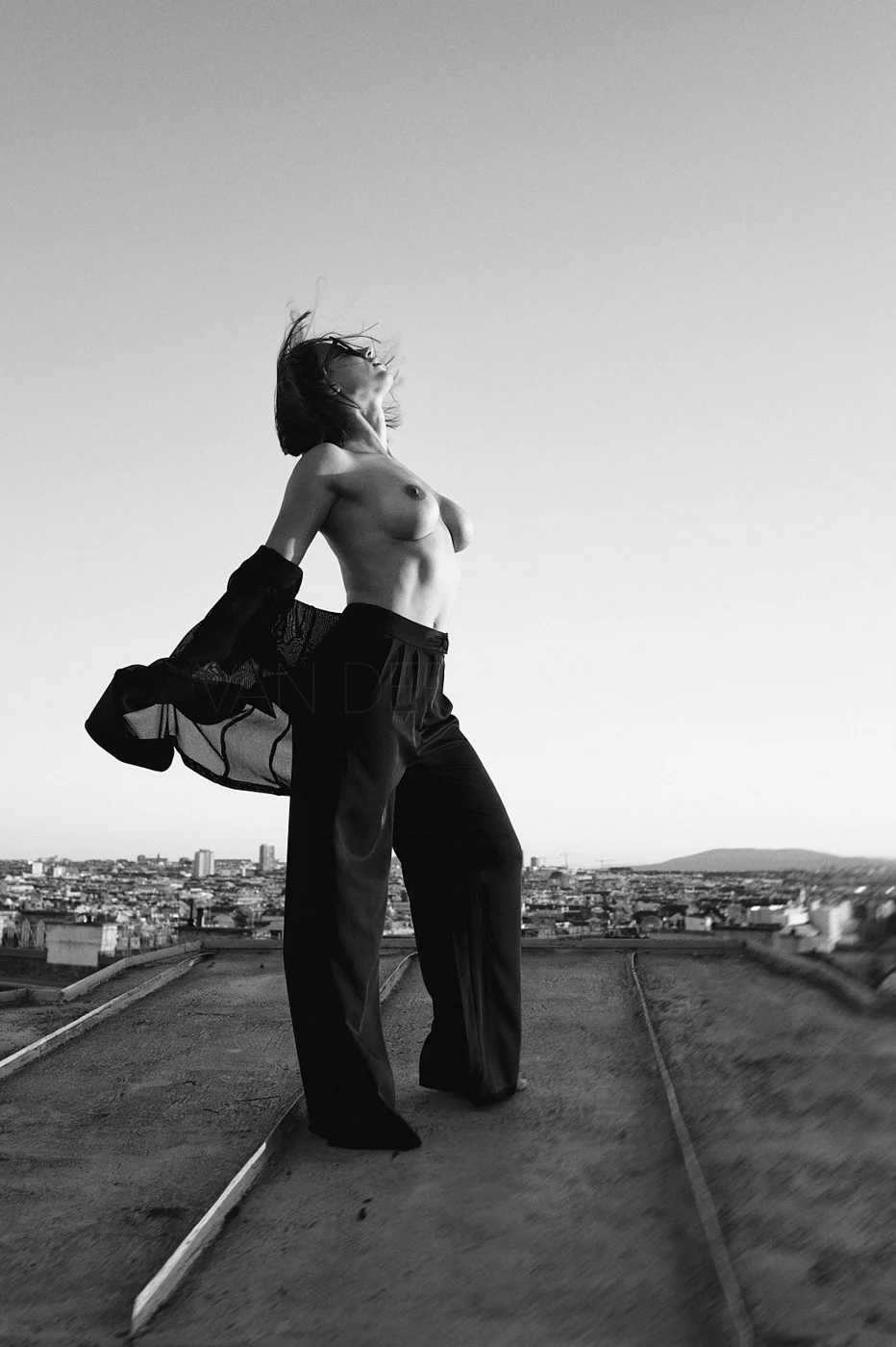 A topless woman standing on a rooftop, wind blowing her hair, with a cityscape in the background, in black and white.