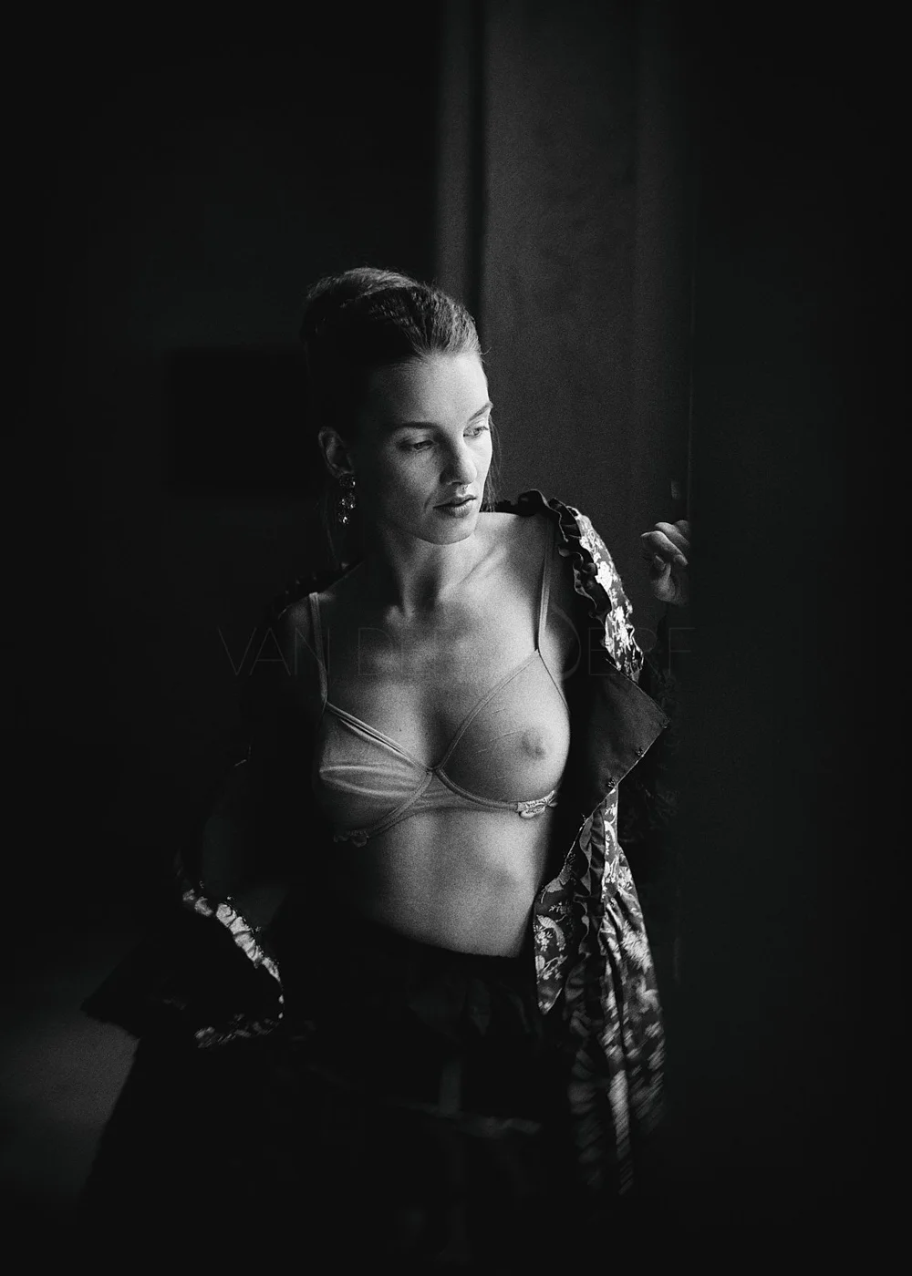 Black and white photo of a woman with her hair in a hairstyle, wearing a transparent bra and a patterned jacket, partially open, standing in a dimly lit environment.
