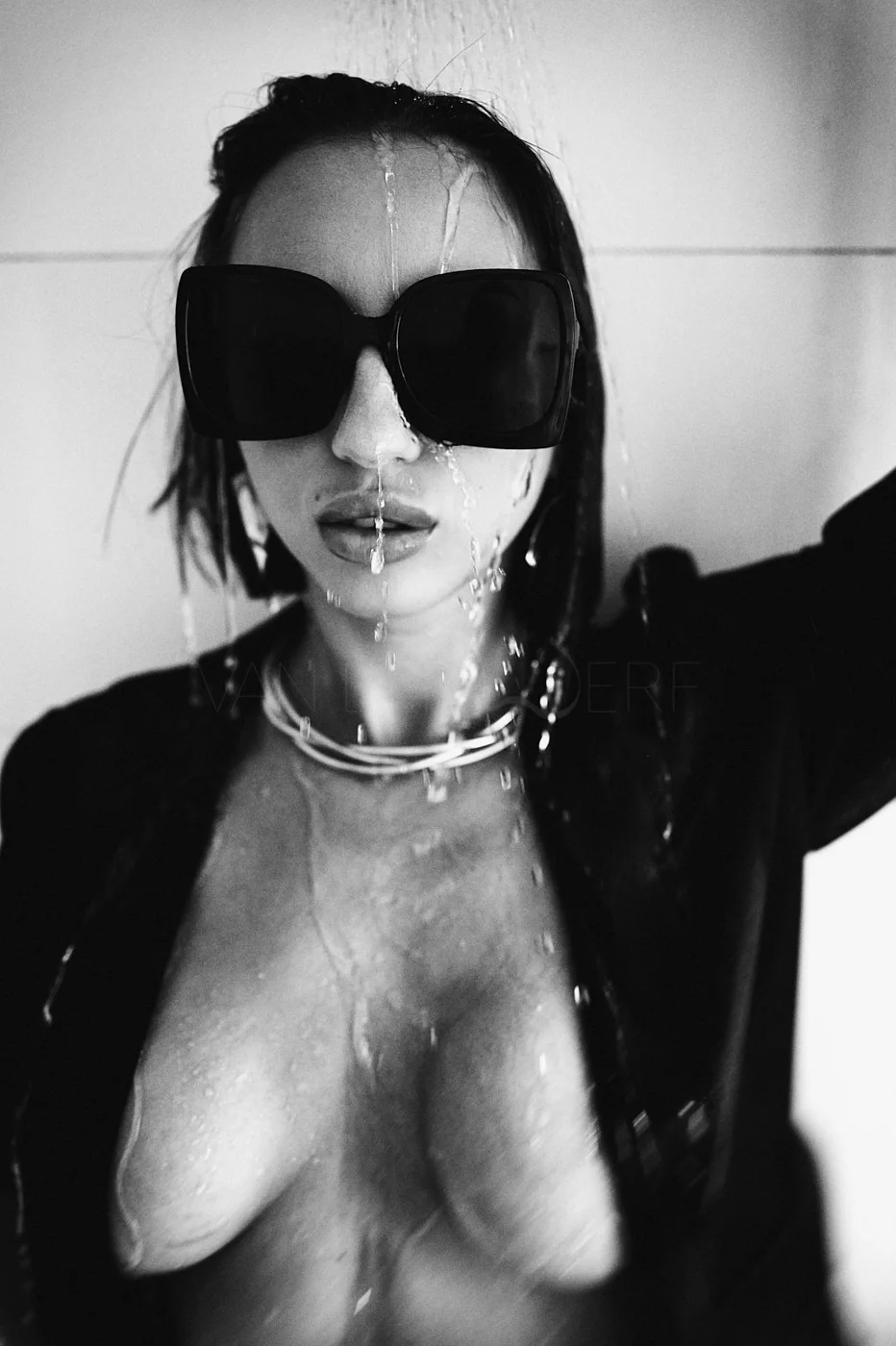 A black-and-white photo of a woman with wet hair, wearing large dark sunglasses, multiple necklaces, and a dark jacket. Her face and chest are wet, with water dripping down.