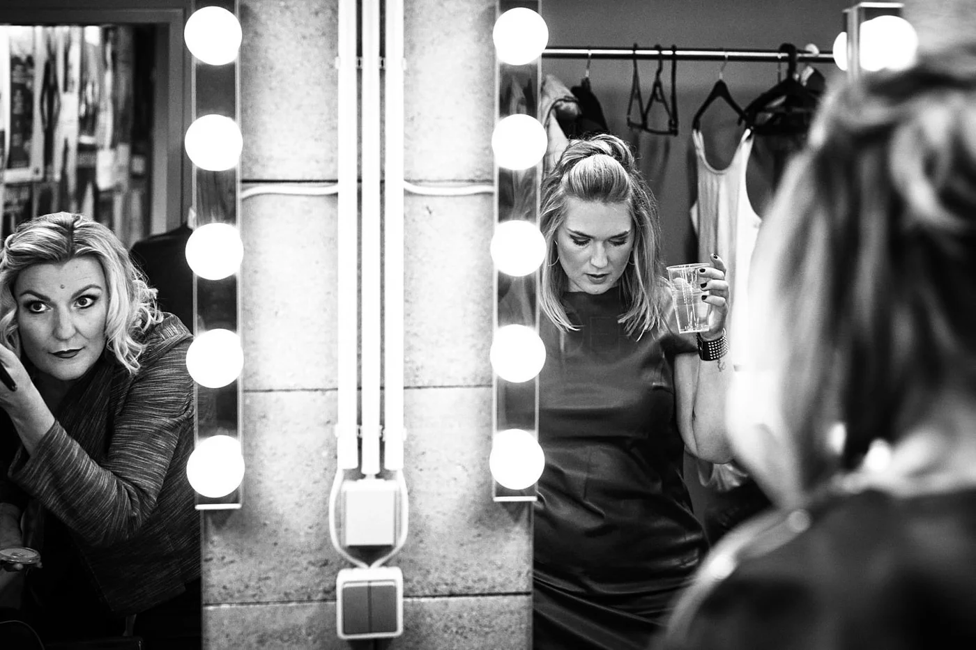 A woman with blonde hair looking into a mirror surrounded by bright light bulbs, applying makeup or styling hair in a dressing room or backstage area.