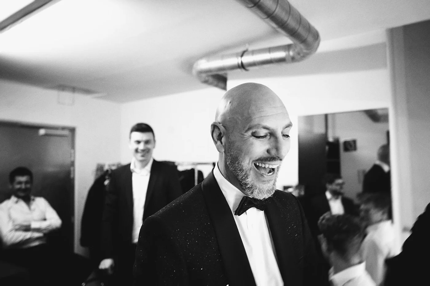 Black and white photo of a smiling bald man with a beard in a tuxedo, at a formal gathering, with people in the background.