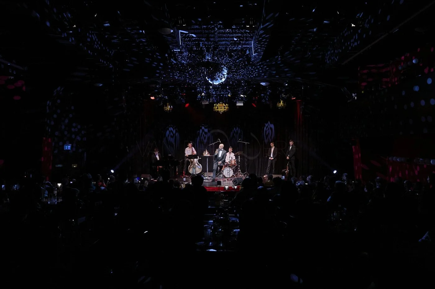A band performing on a dimly lit stage with a backdrop of reflective disco balls casting dots of light, and an audience seated at tables.