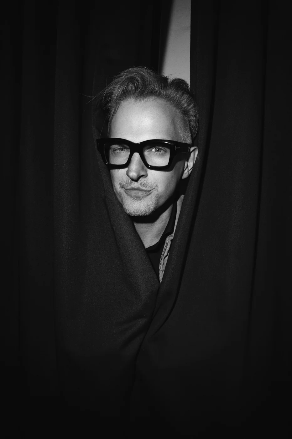Black and white portrait of a man with short, styled hair wearing large, thick-rimmed glasses, peeking through black curtains.