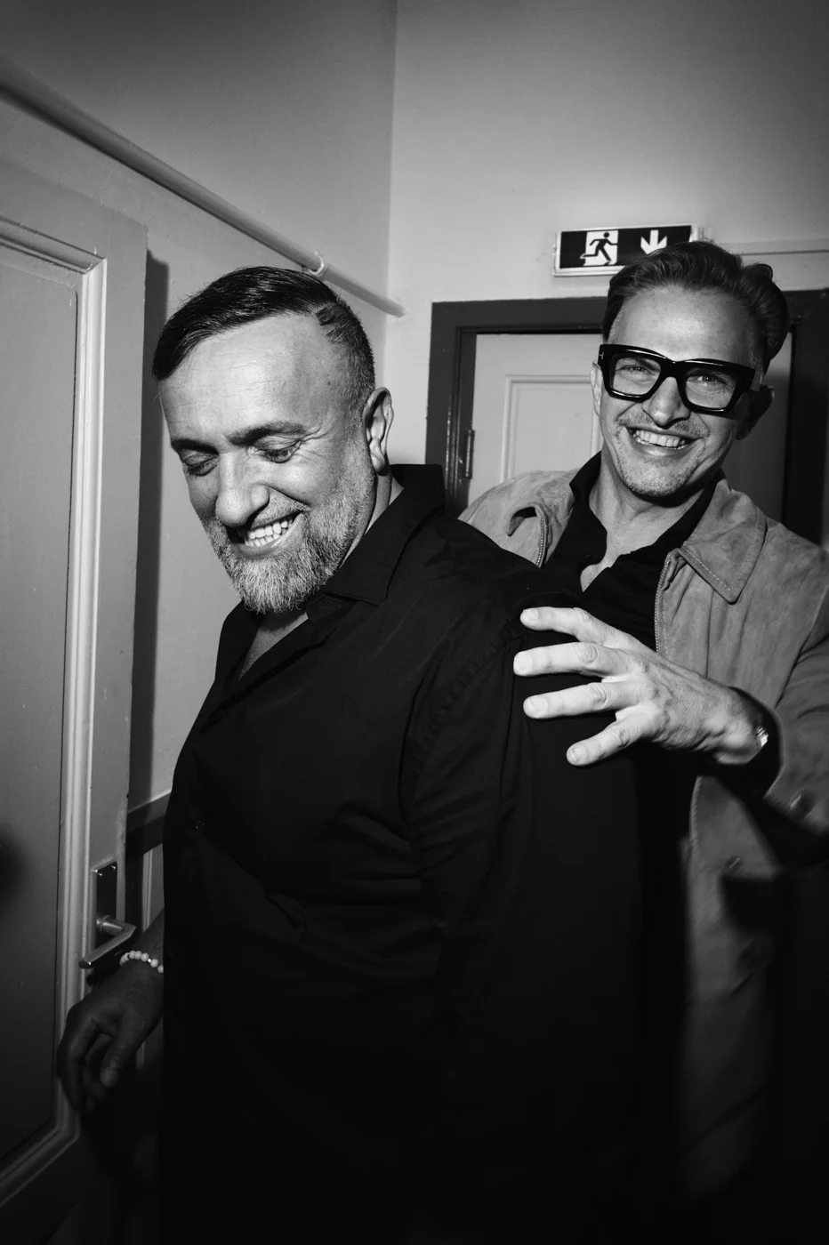 Two men smiling and playfully interacting indoors with one man standing behind the other, who is holding a door handle.