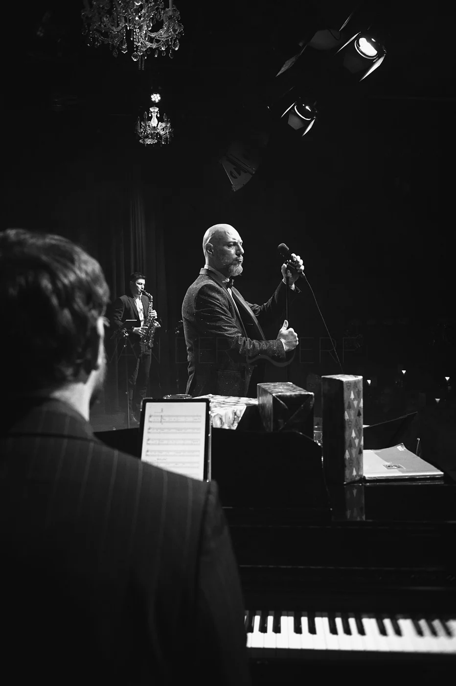 Black and white photo of a jazz performance on stage. A man in a suit is holding a microphone, singing, with a saxophonist and a pianist in the background. There are chandeliers hanging from the ceiling.