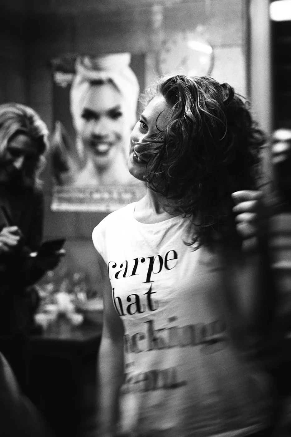 A woman with curly hair is flipping her hair in a hair salon, wearing a white t-shirt with text, and smiling. In the background, there is a poster of a woman with blonde hair.