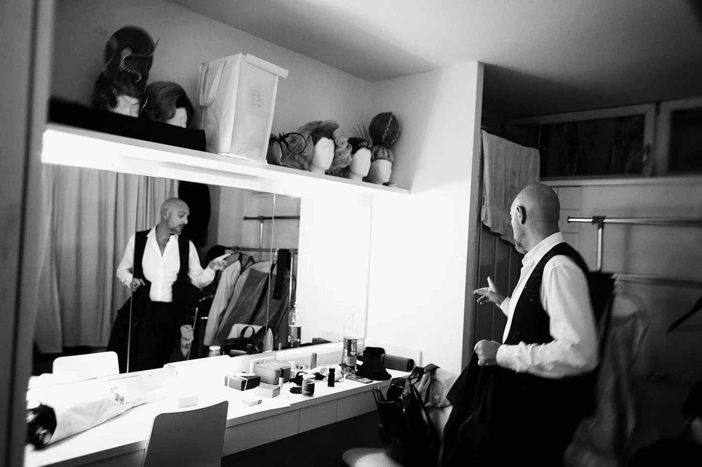 A man in formal attire is sitting in a dressing room, looking at his phone. Another man in similar attire is standing in front of a mirror, adjusting his clothing. The room has a cluttered white counter with various items, and shelves above with mann