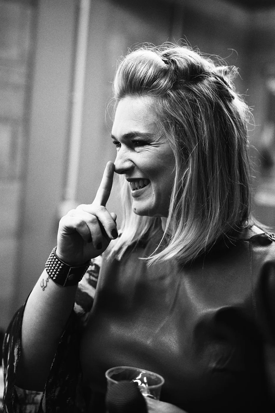 Black and white photo of smiling woman with shoulder-length hair, wearing a leather jacket and studded bracelet, holding a drink, with her finger near her nose.