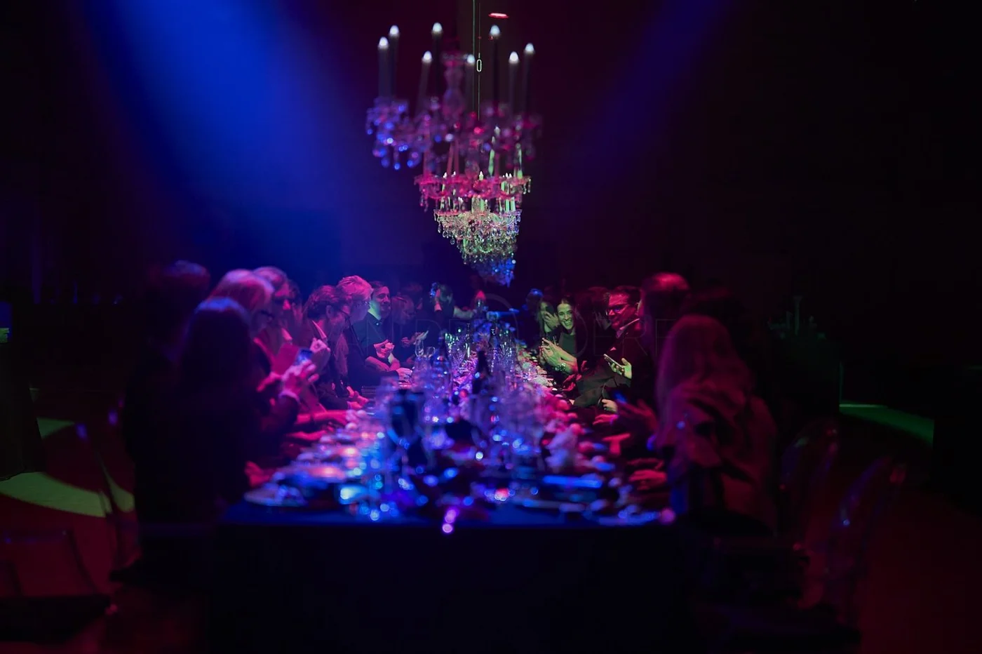 People sitting at a long dinner table with colorful lighting, a chandelier hanging above, in a dark room during an event or celebration.