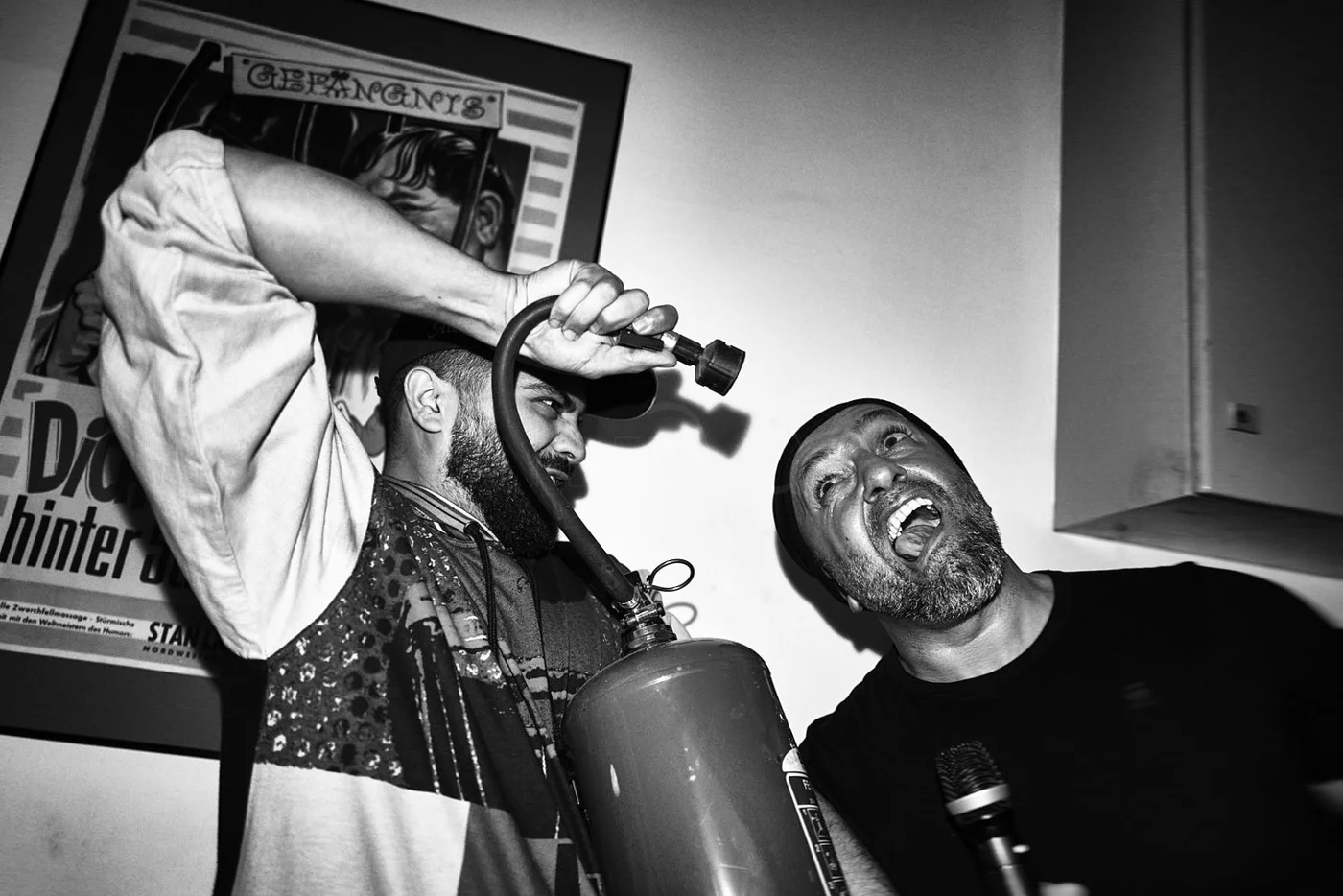 Two men with facial hair in a black and white photo, one holding a fire extinguisher and aiming it at the other's face, who appears to be shouting. There is a poster on the wall behind them.