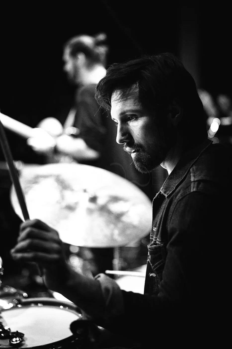 Black and white photo of a man with dark hair and beard playing drums, with a woman in the background also playing drums, both in focused mood.