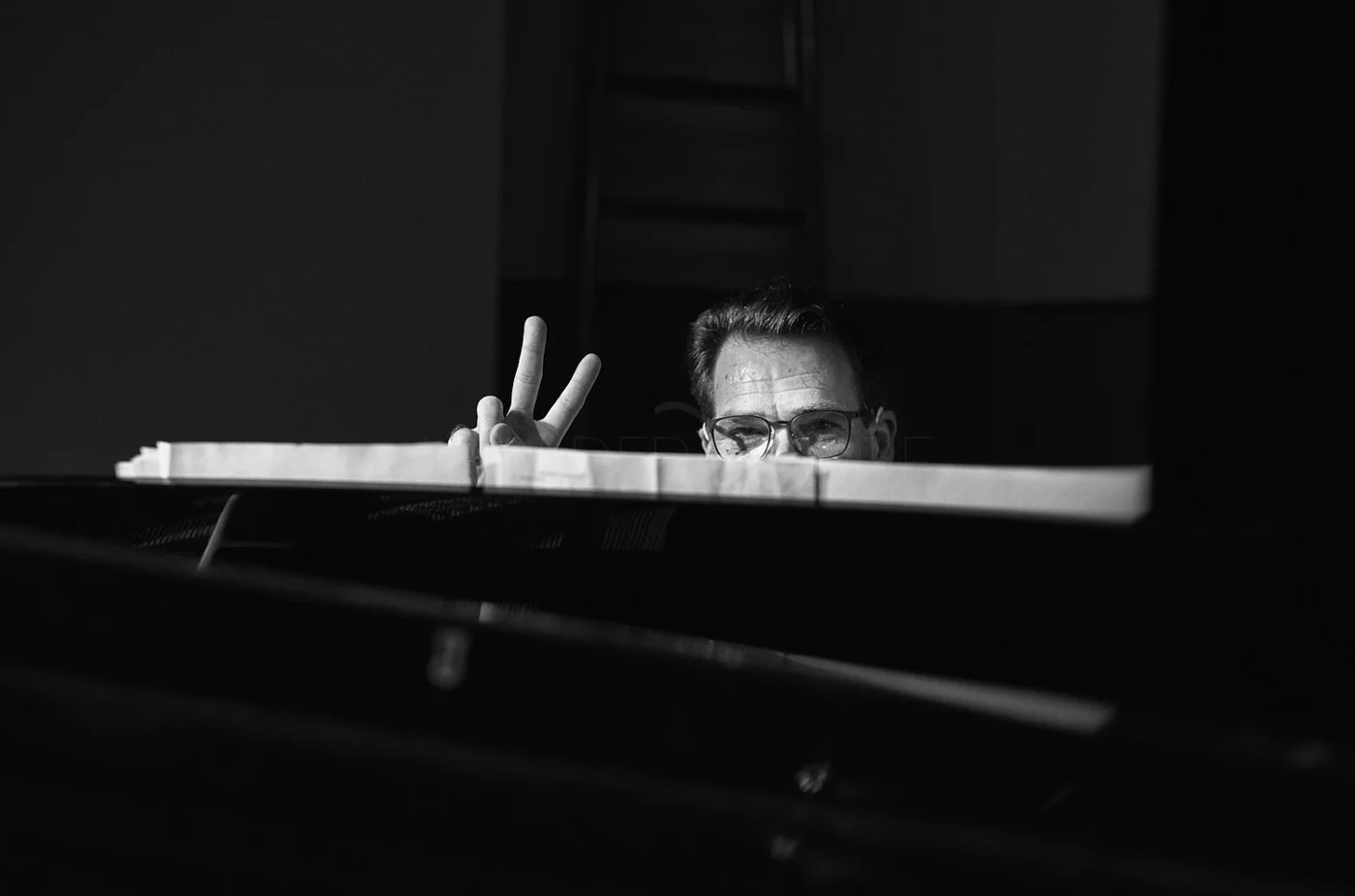 A person with glasses makes a peace sign behind a piano, smiling, in black and white.