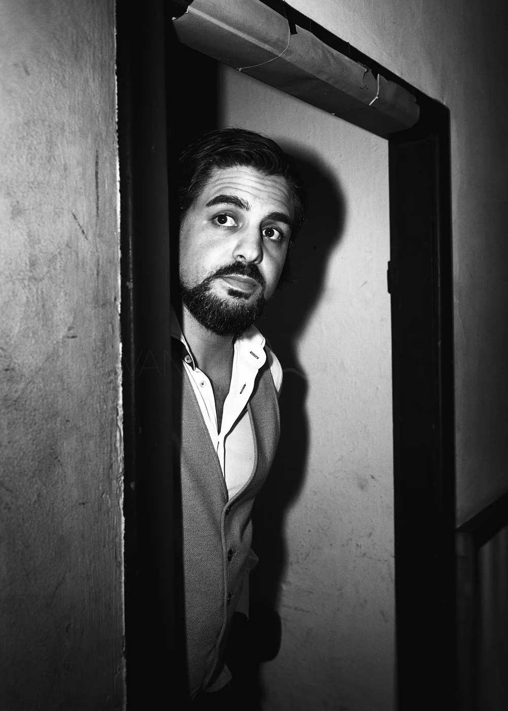 A man with dark hair and a beard peeks through a doorway, looking directly at the camera in black and white.