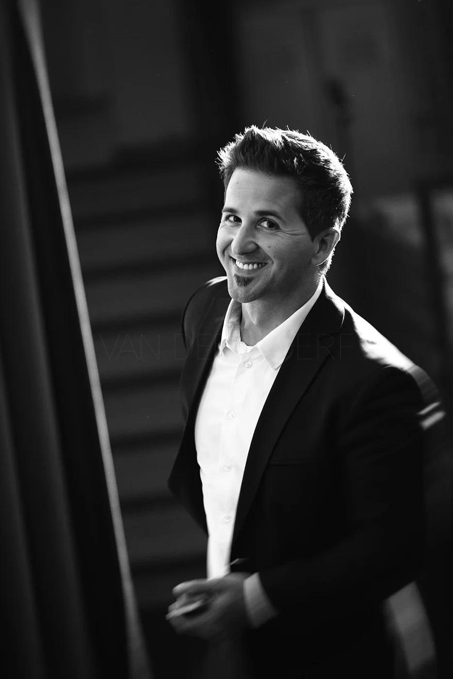 A black and white portrait of a smiling man in a suit and white shirt, looking at the camera.