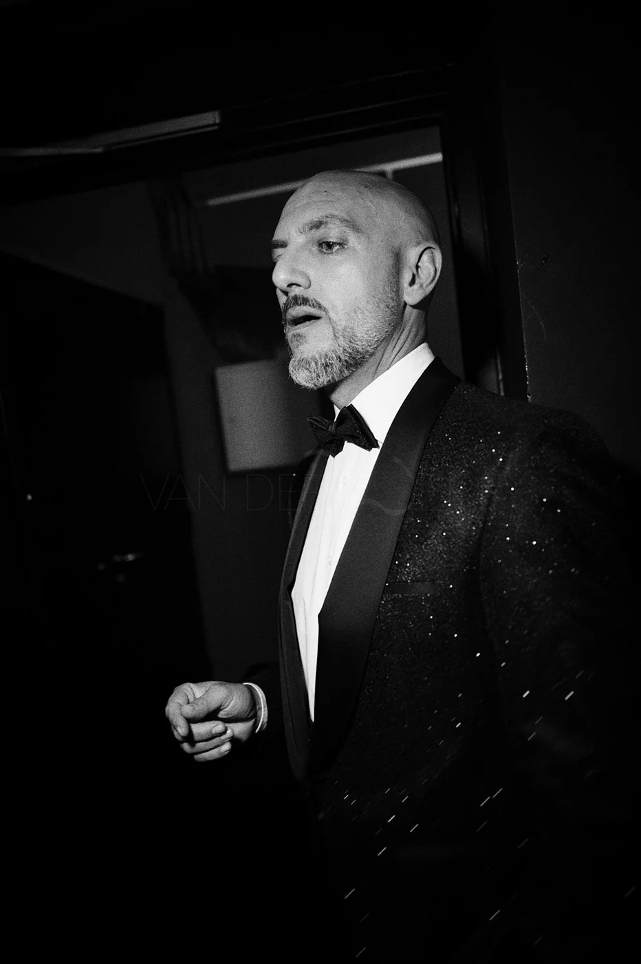 A black and white photo of a man in a tuxedo, walking with a serious expression, in a dimly lit environment.