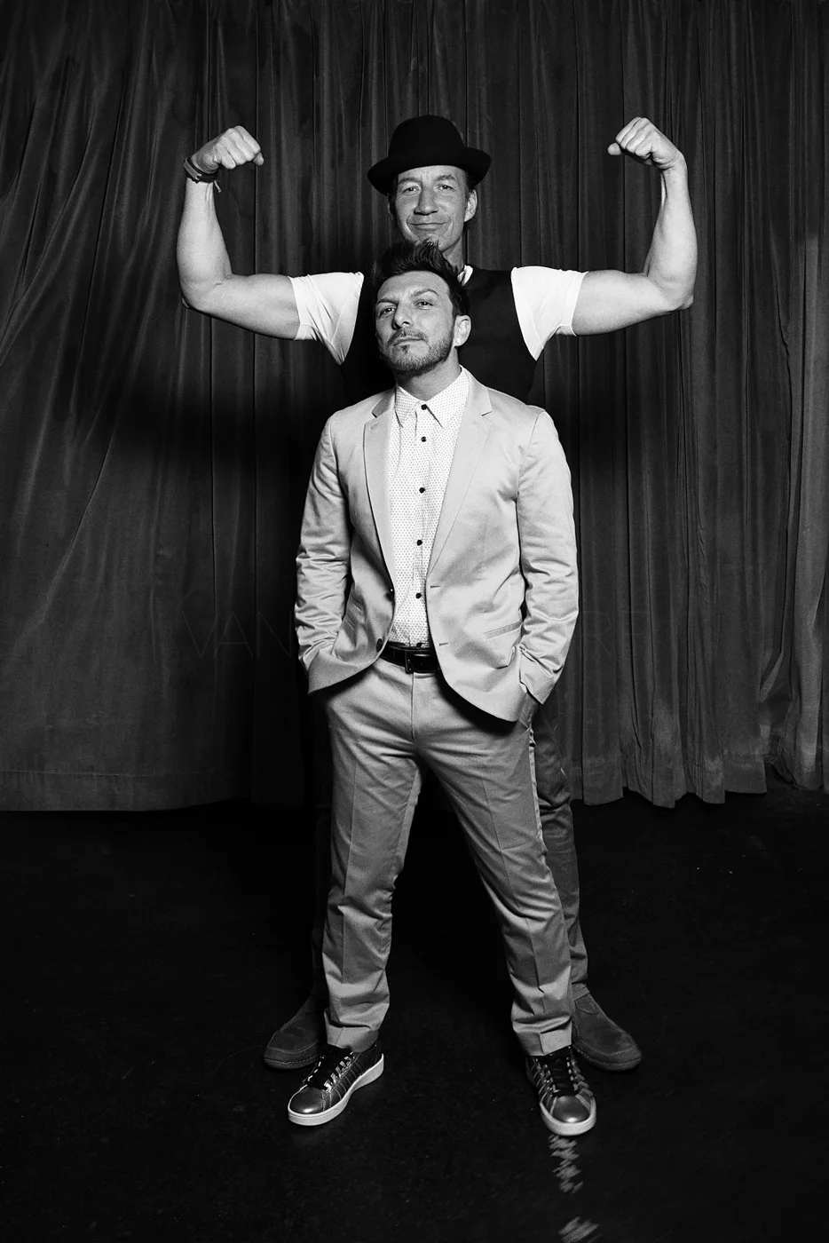 Two men, one tall with a bowler hat and flexed arms, standing behind a shorter man in a suit, all in front of a curtain.