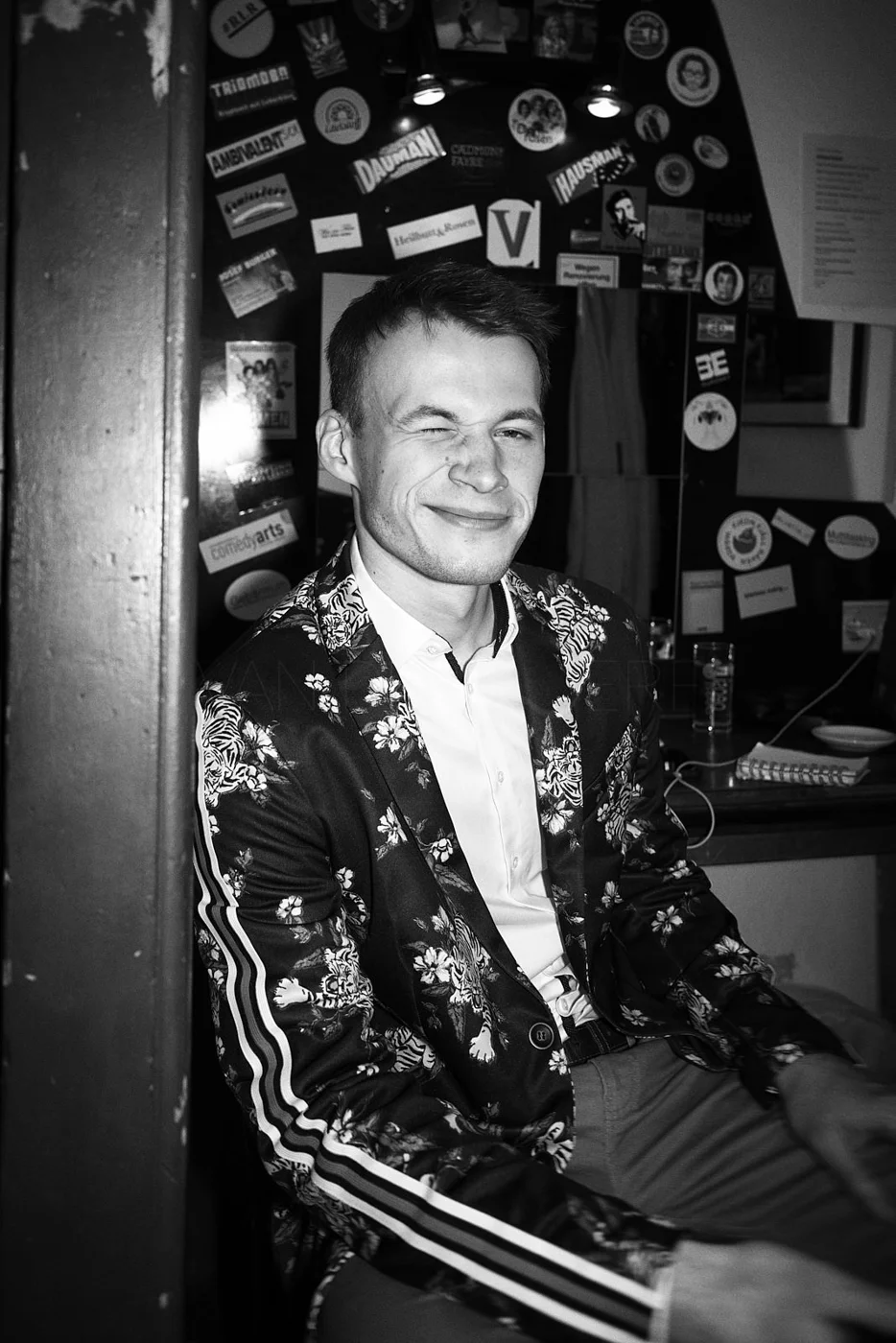 A young man winking and smiling at the camera, sitting in a room with various stickers and posters on the wall behind him, wearing a floral-patterned jacket over a white shirt.
