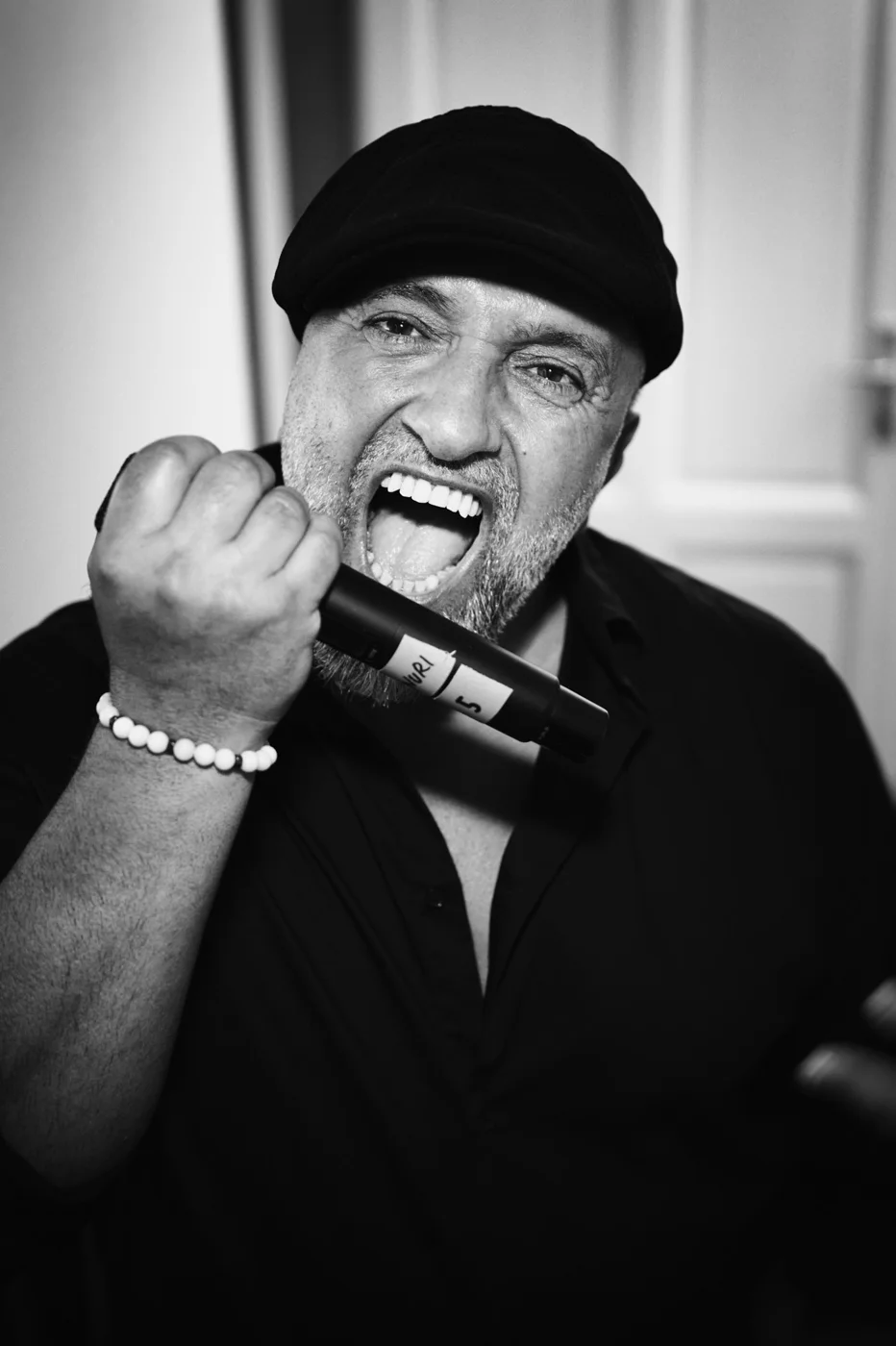 A man with a beard and wearing a black cap, passionately singing or shouting into a microphone, with a wide open mouth and expressive eyes, in a black and white photo.