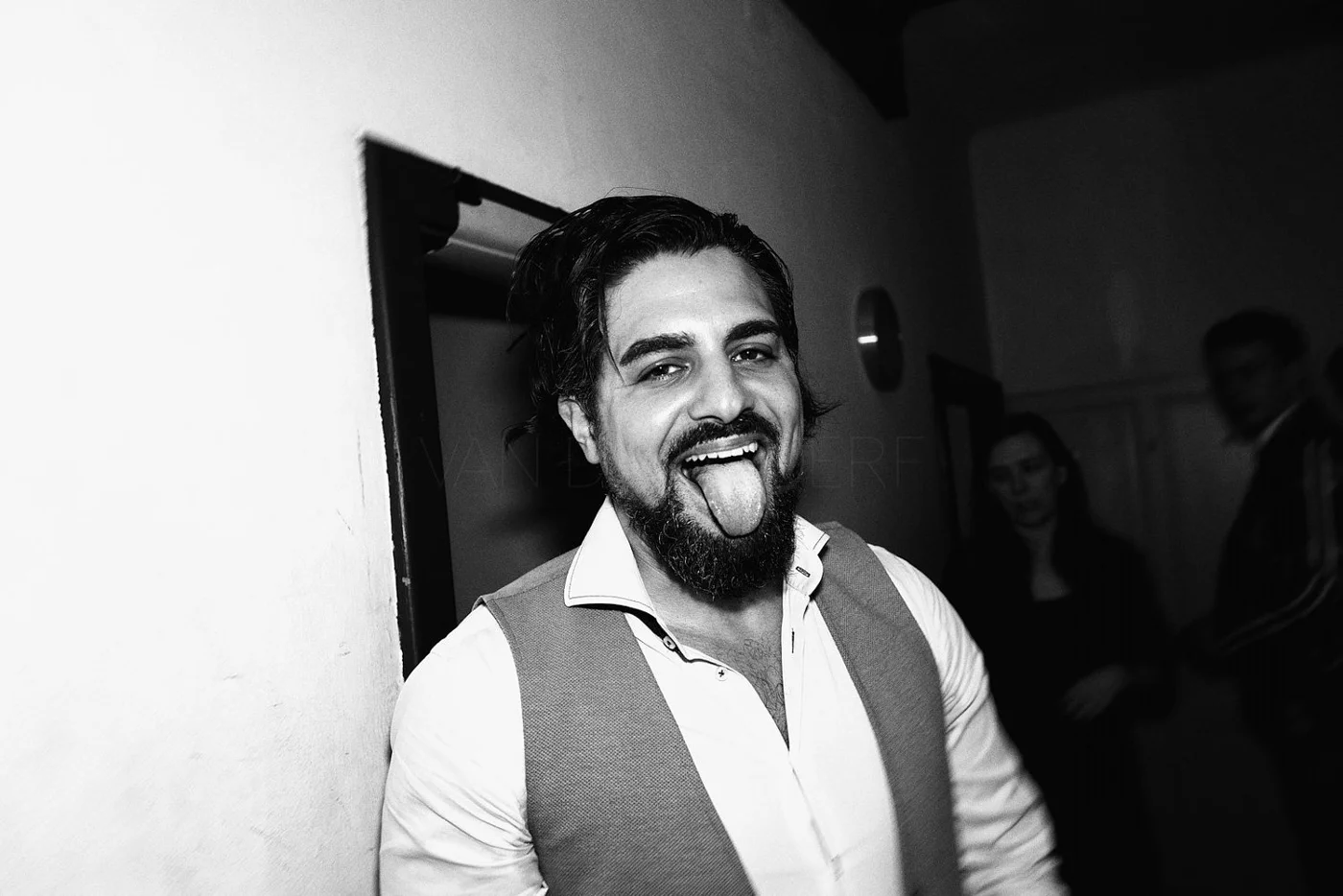 A man with dark hair and a beard, wearing a light-colored shirt and vest, sticking out his tongue, smiling, in an indoor setting.
