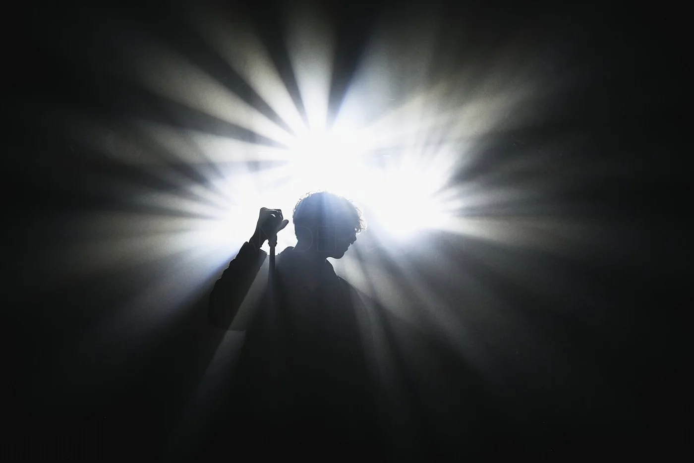 Silhouette of a woman with short curly hair standing in front of a bright light with rays radiating outward, creating a dramatic silhouette effect.