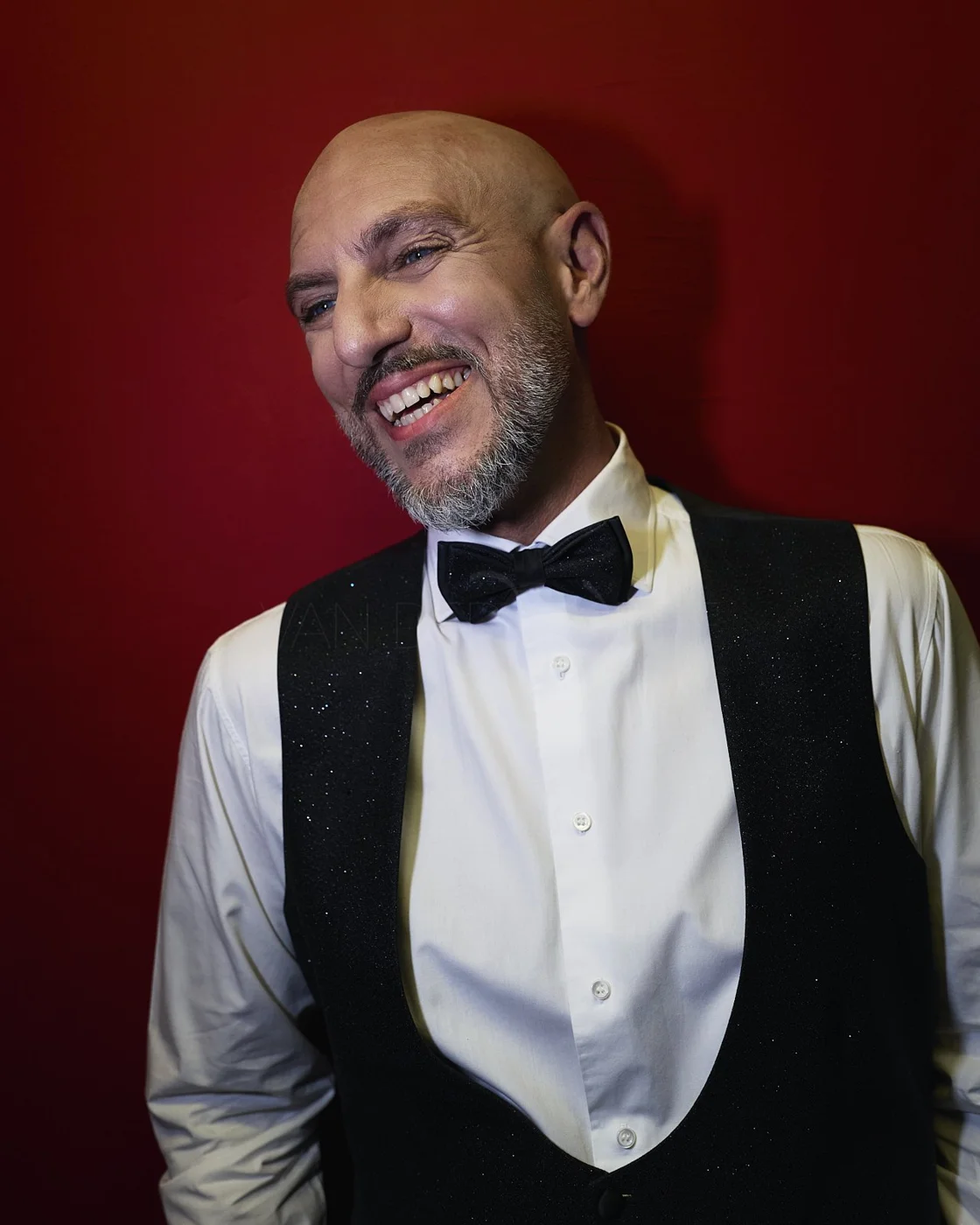A smiling man in a tuxedo with a black bow tie and black vest, standing against a red background.