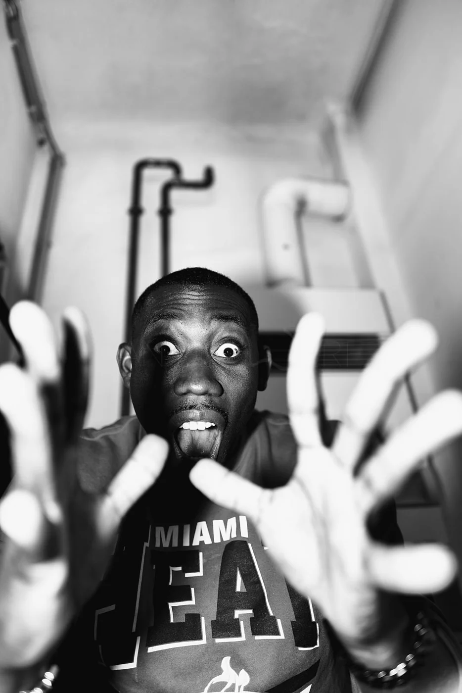 A man with wide eyes and tongue out, making a playful face, reaching towards the camera with both hands in front of him. The background shows pipes and an air conditioning unit.