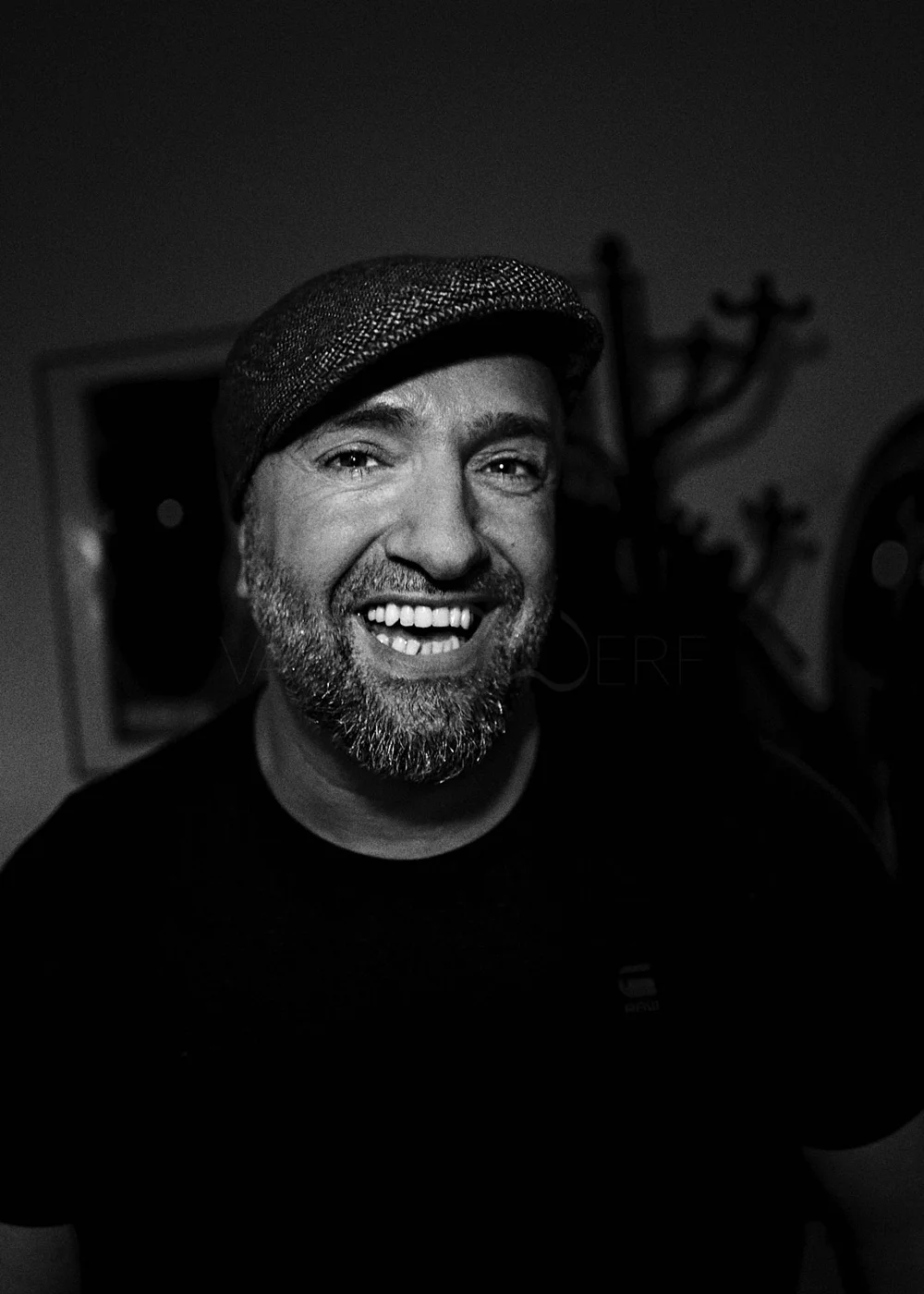 Black and white photo of a smiling man wearing a flat cap and a dark shirt, with a blurred background featuring abstract artwork and a coat rack.