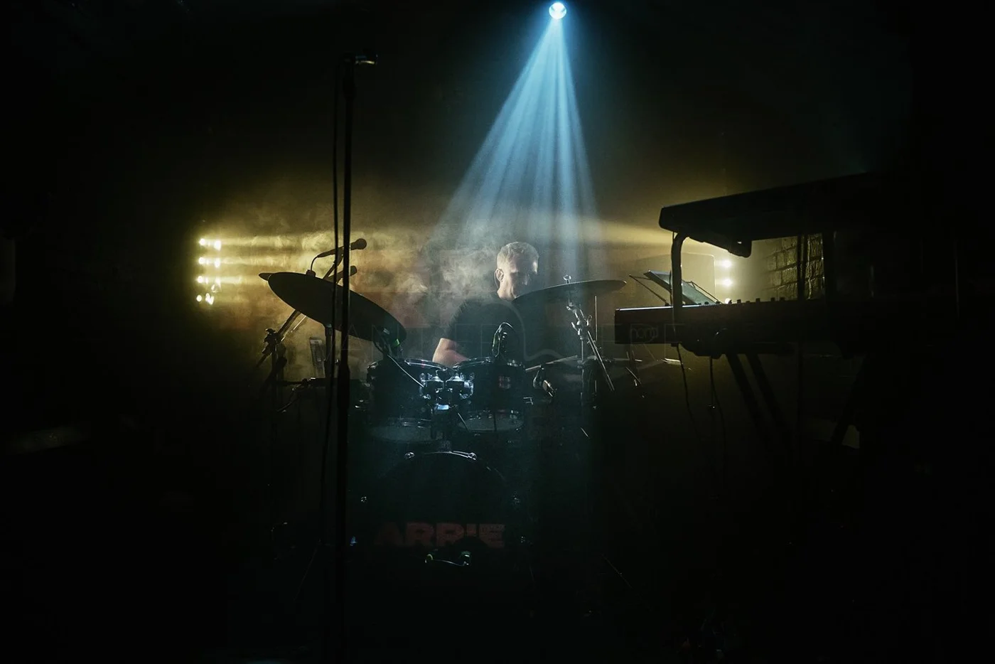 A musician playing the drums on stage with colorful stage lights shining down, surrounded by musical equipment.