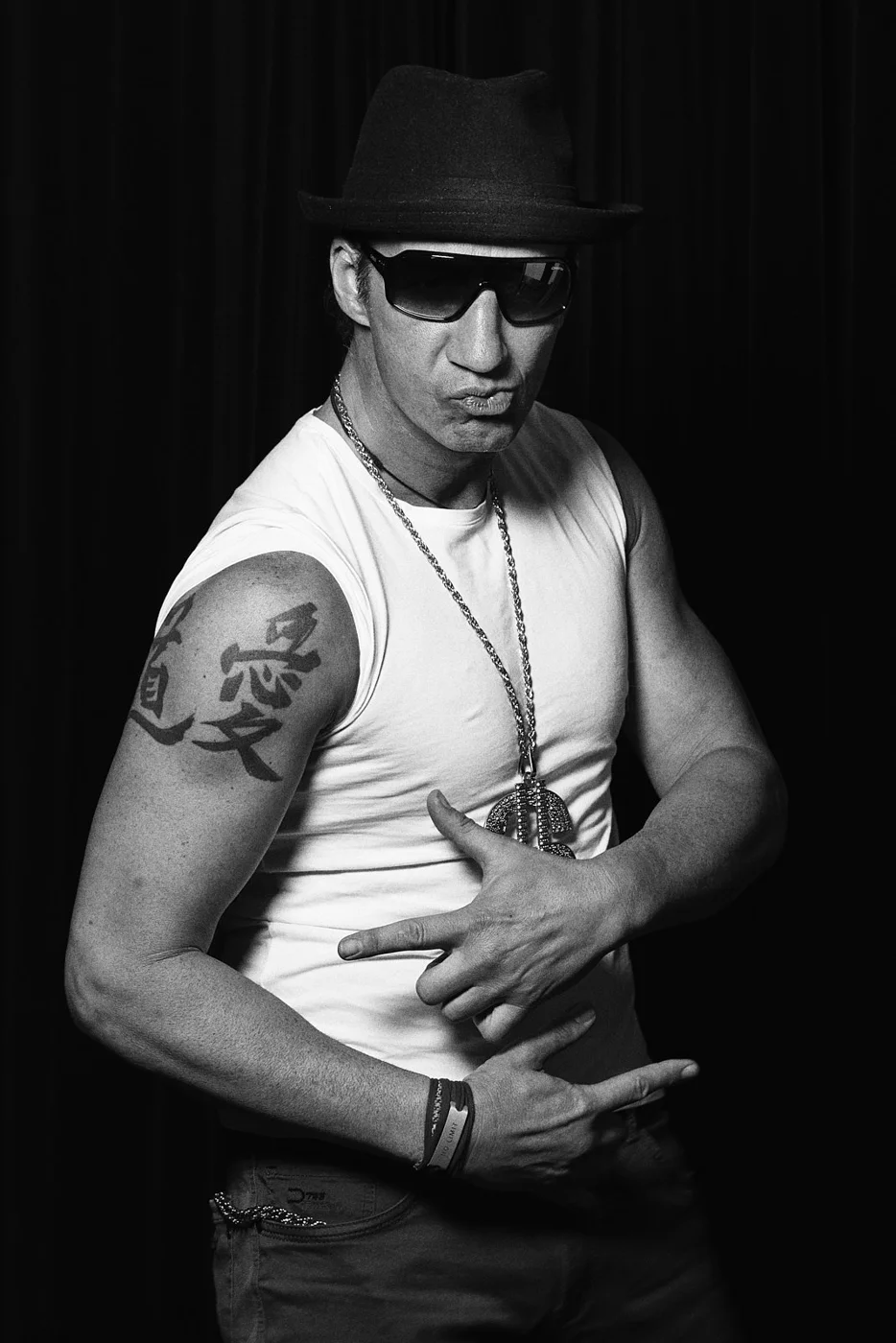 A man wearing a hat and sunglasses making a hand gesture, with tattoos on his arm and a large chain necklace, standing against a dark background.