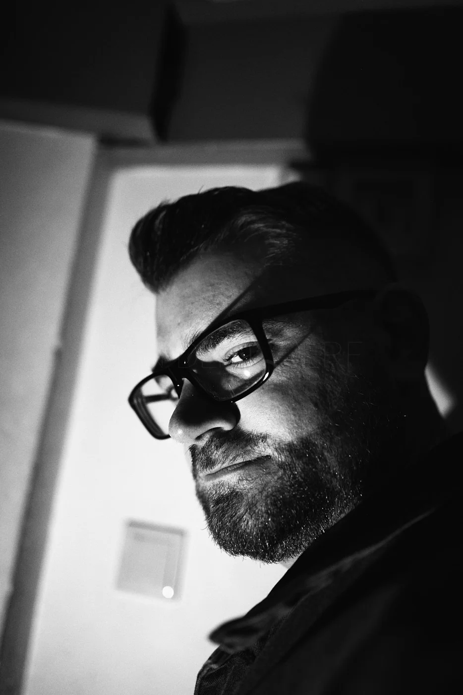 Black and white close-up portrait of a man with glasses, beard, and slicked-back hair, looking over his shoulder.