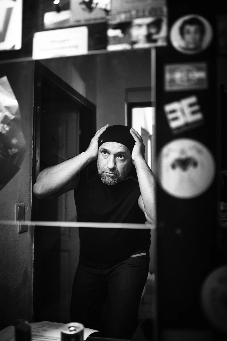 A man looks at himself in a mirror, adjusting a beanie, with cluttered stickers and photos on the wall behind him.