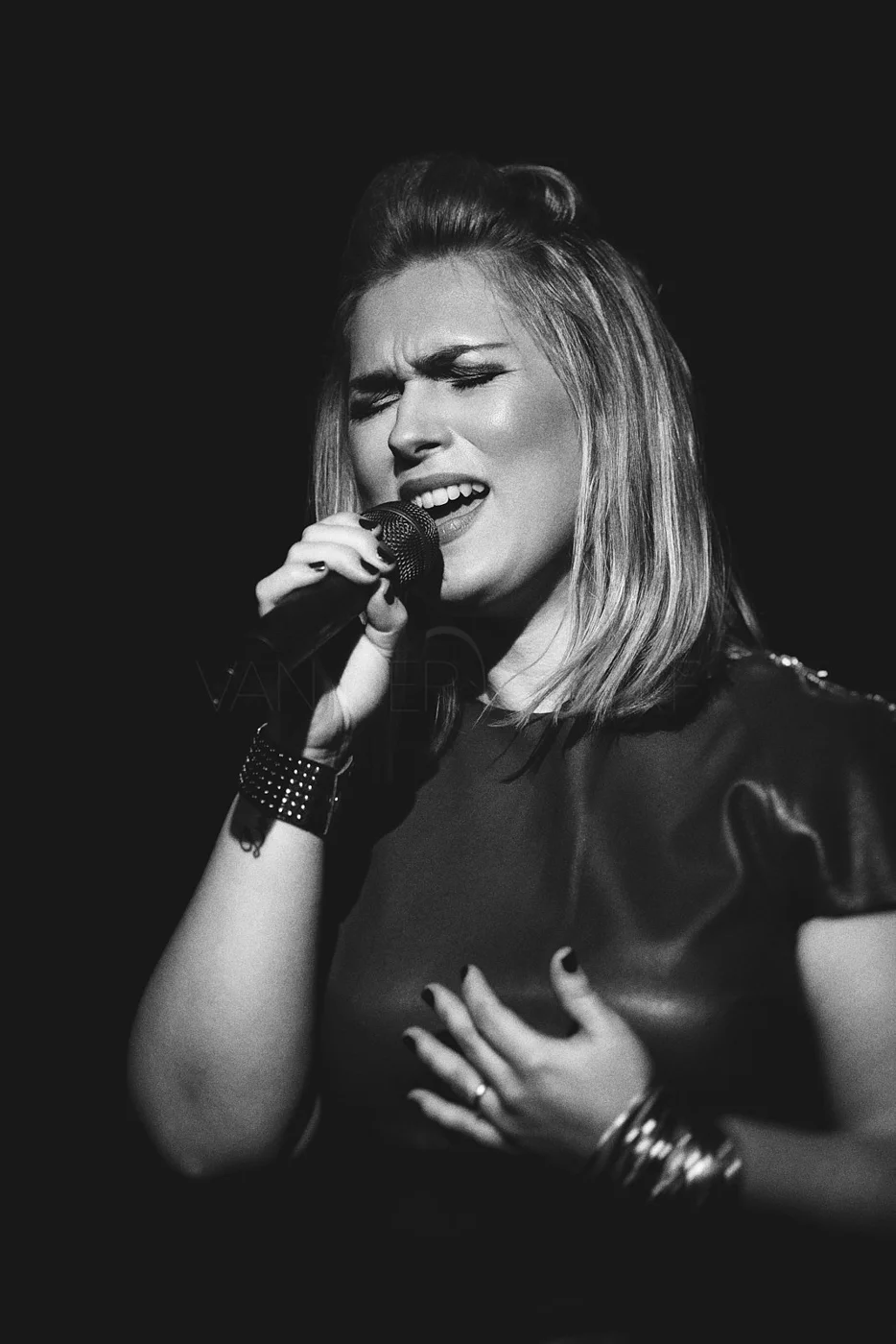 A woman with blonde hair singing passionately into a microphone, eyes closed, wearing a dark top and bracelets, in a black-and-white photo.
