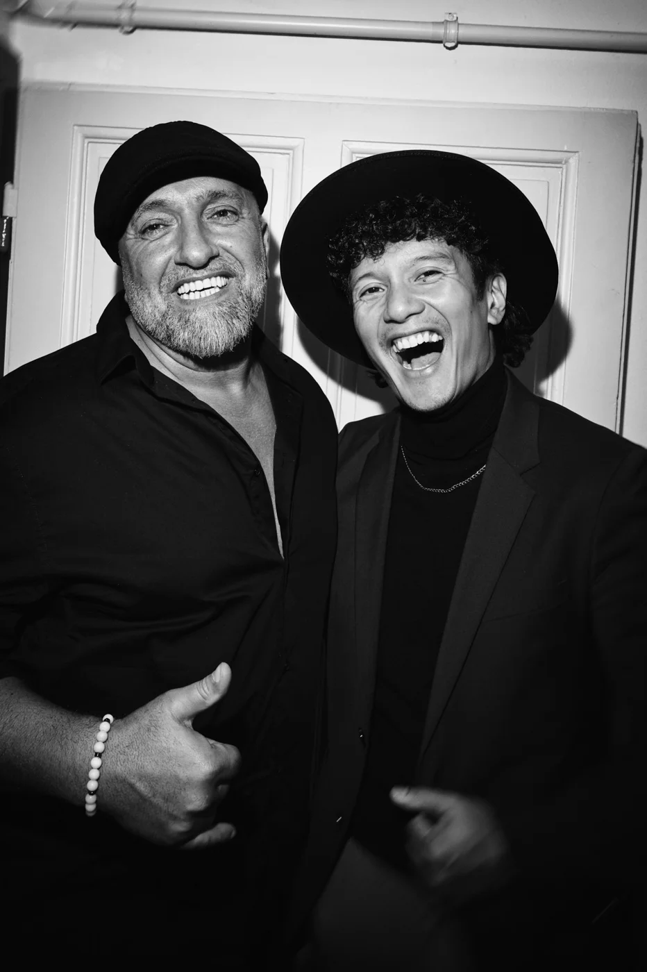 Two men smiling and posing together indoors, one giving a thumbs-up and the other showing a fist bump, both wearing dark clothing and hats.
