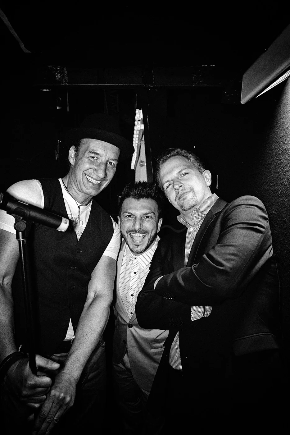Three men smiling and posing for a photo in a dark setting, one holding a microphone, one with crossed arms, all dressed in semi-formal to formal attire.