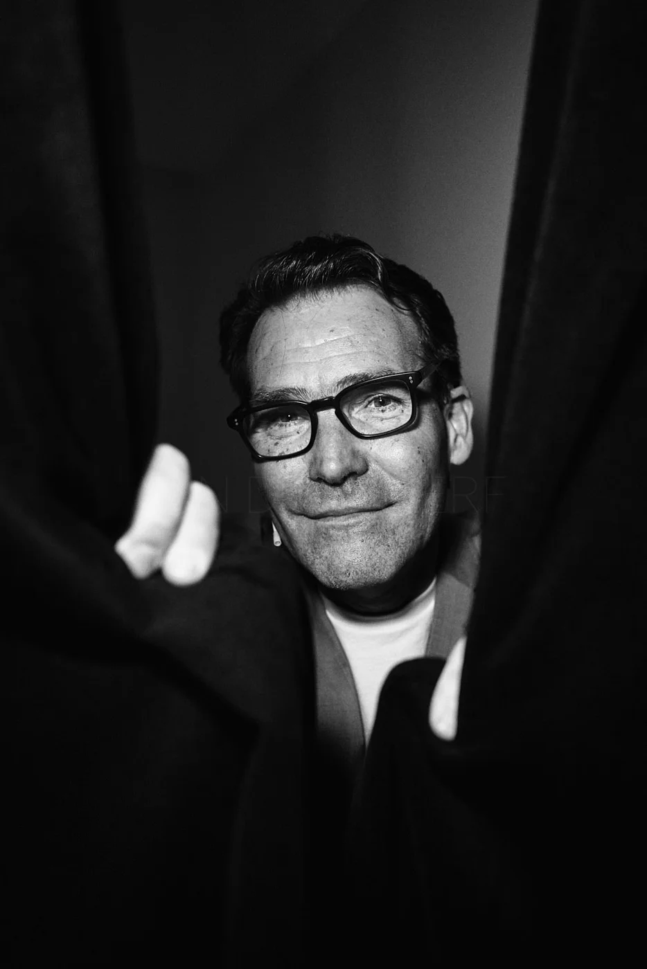 Black and white photo of a man with glasses looking into the camera, smiling, with two people partially visible in the foreground.