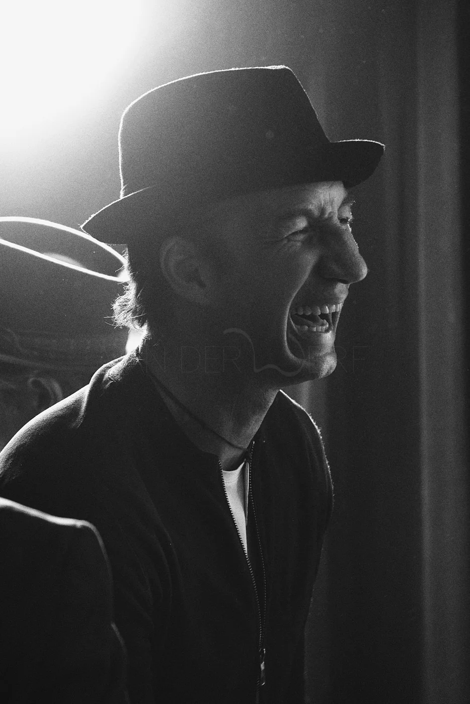 A man wearing a hat and jacket, laughing with his eyes closed, in black and white.