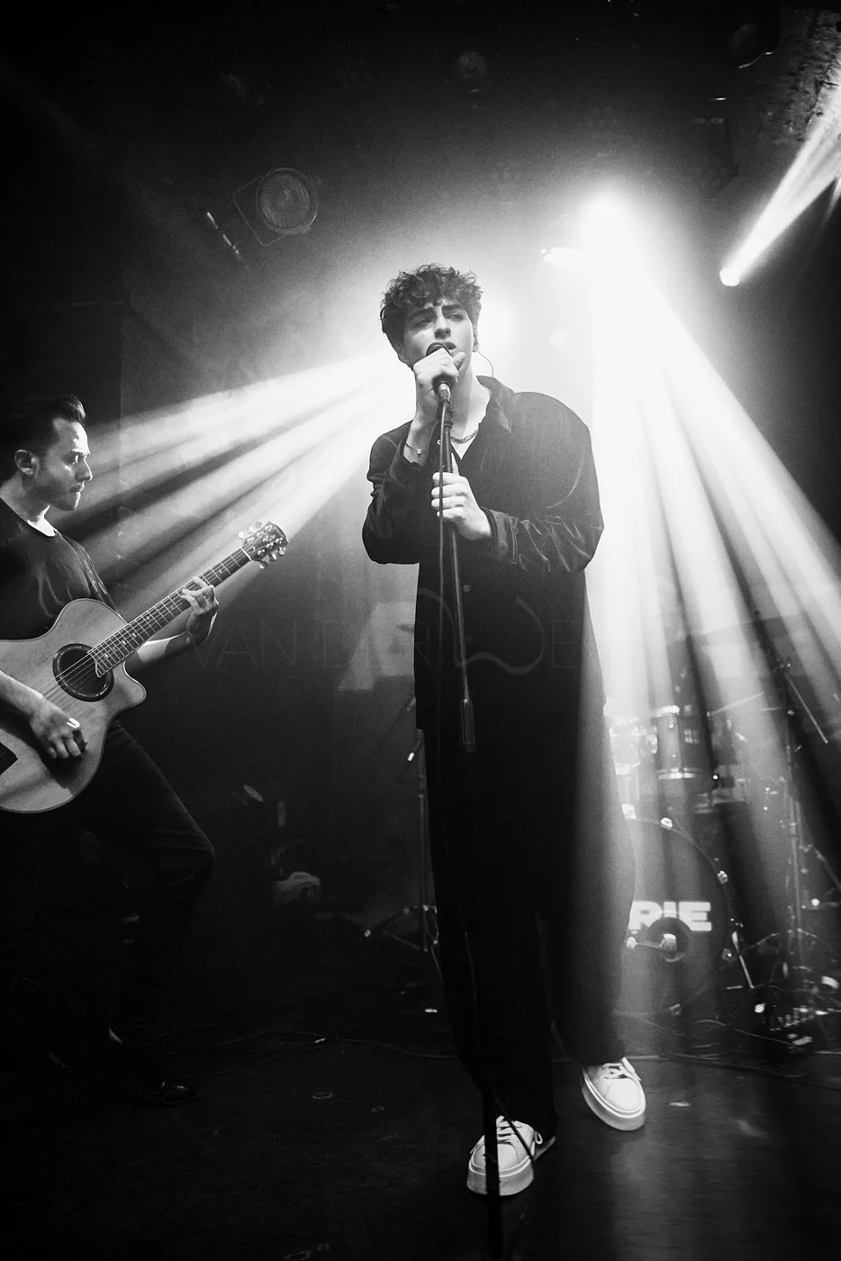 Black and white image of a male singer performing with a microphone, accompanied by a guitarist on stage, with stage lights creating beams of light.
