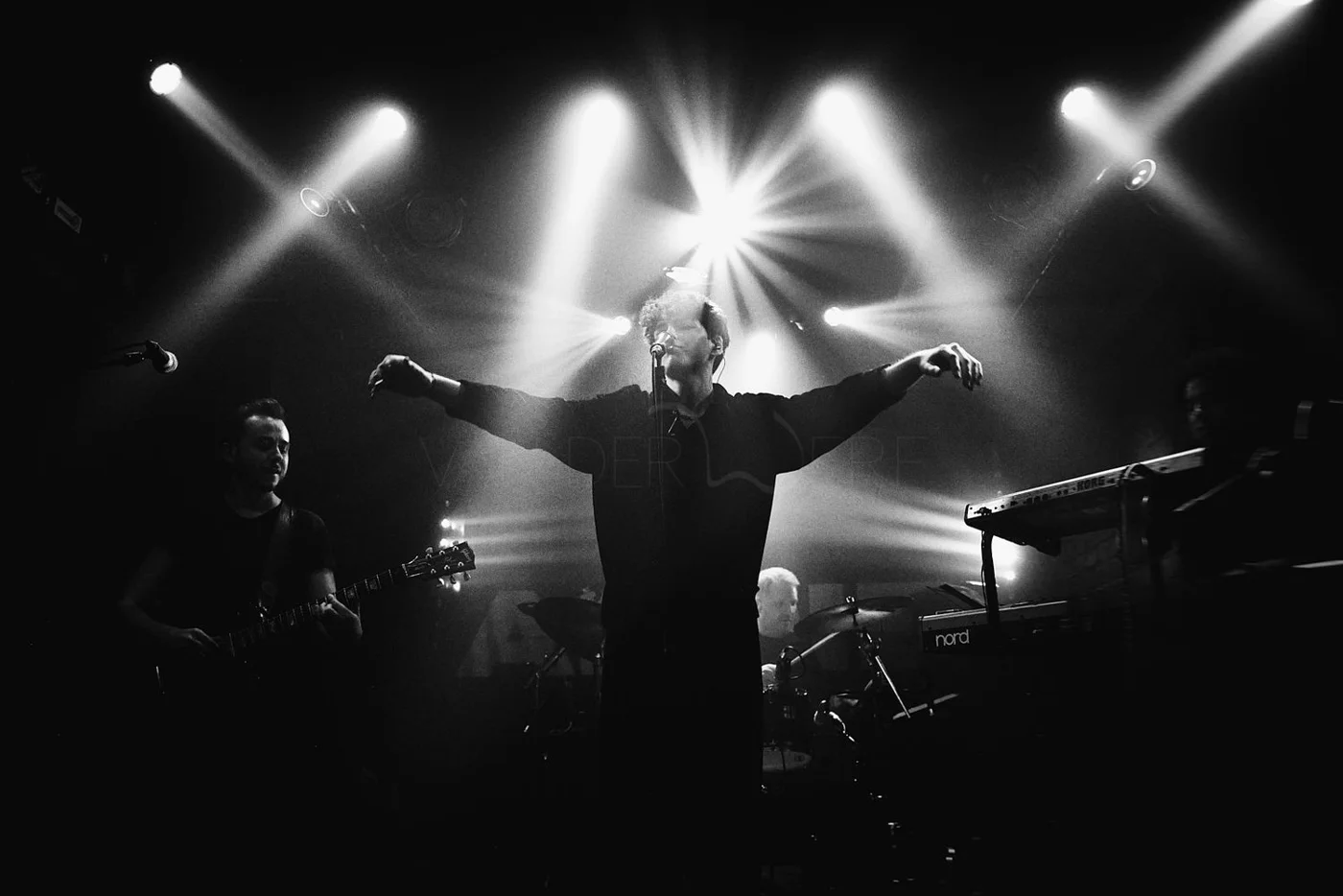 A black and white photo of a band performing on stage with a singer in the center with arms outstretched and lights shining behind them.