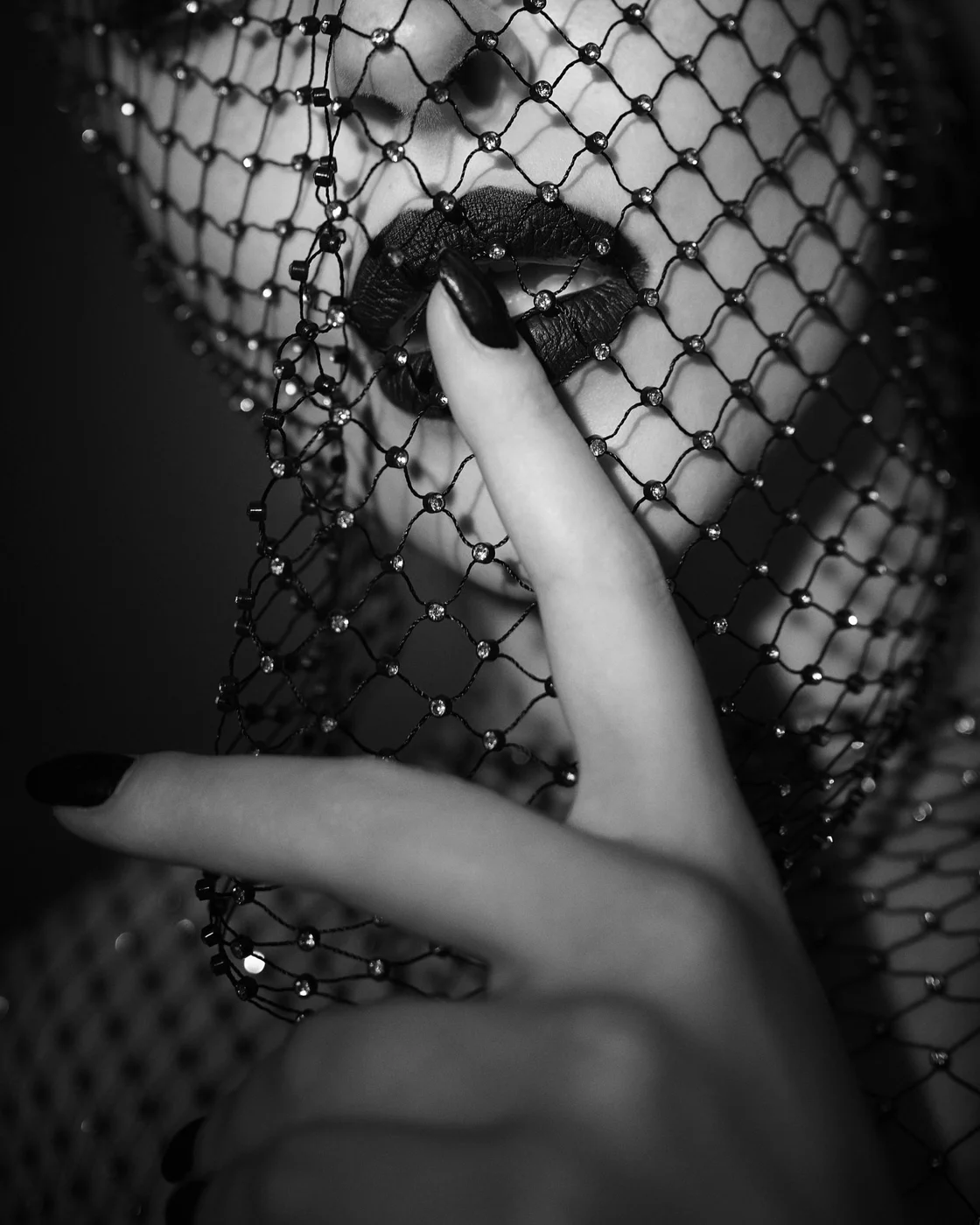 Black and white photo of a woman's face partially covered by a net with gems, showcasing dark lipstick and a finger over lips.
