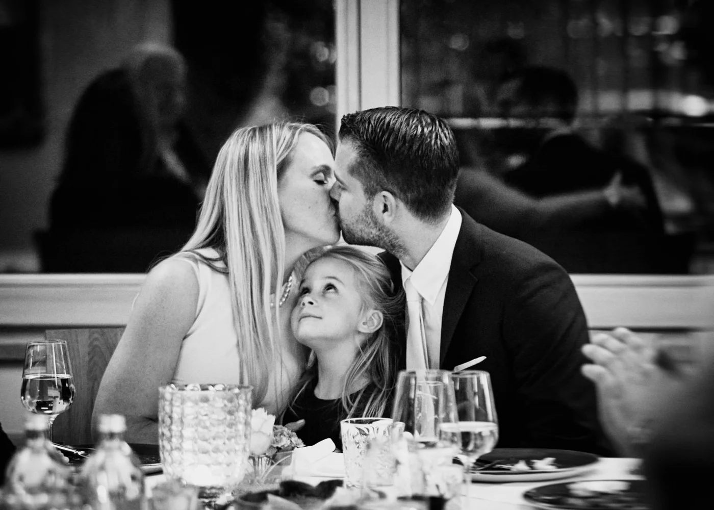 A woman and a man are kissing at a dinner table while a young girl looks up at them, with glasses and tableware in front of them.