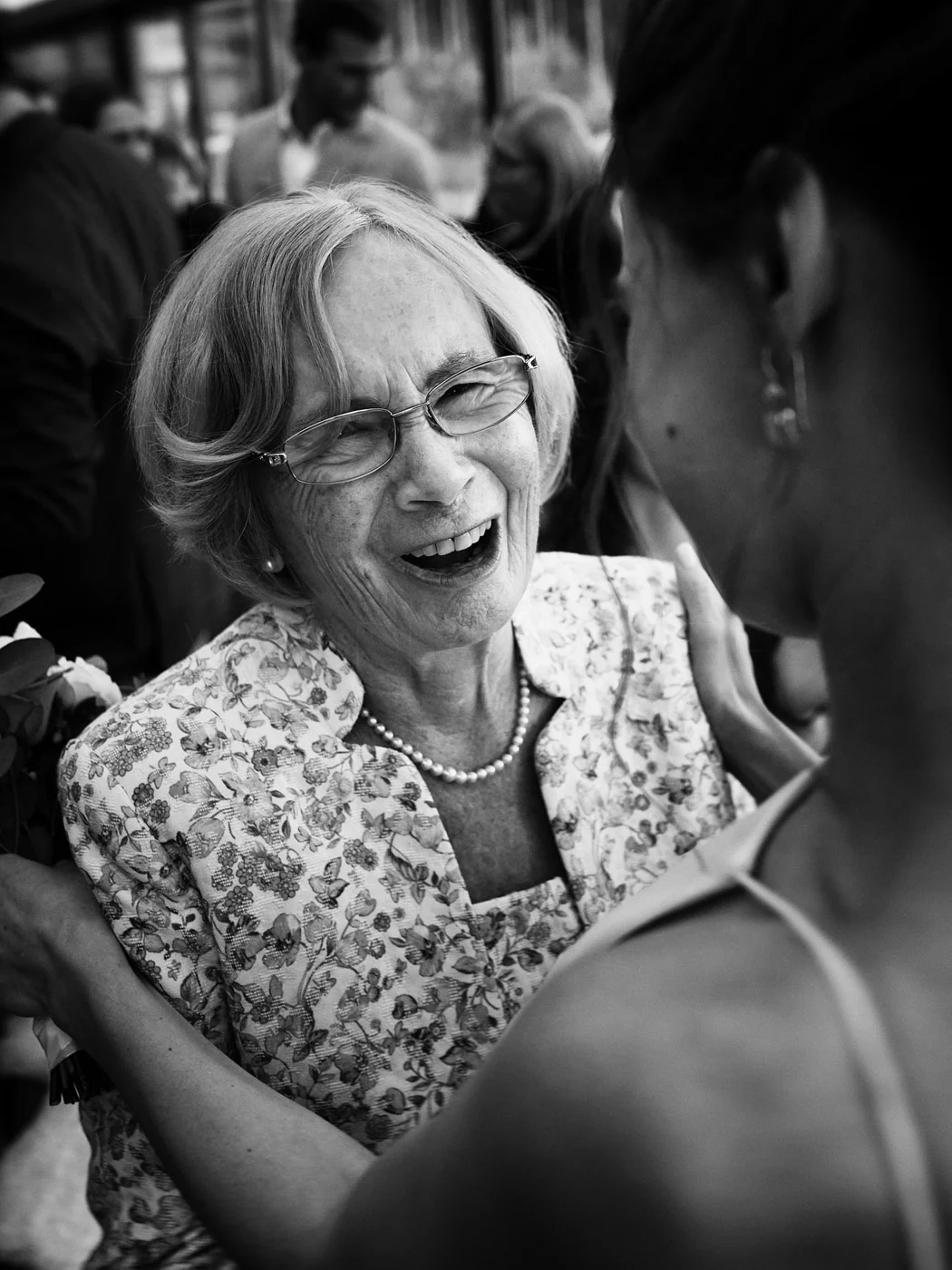 An elderly woman with glasses and a floral blouse smiling as she embraces a younger woman during a social gathering.