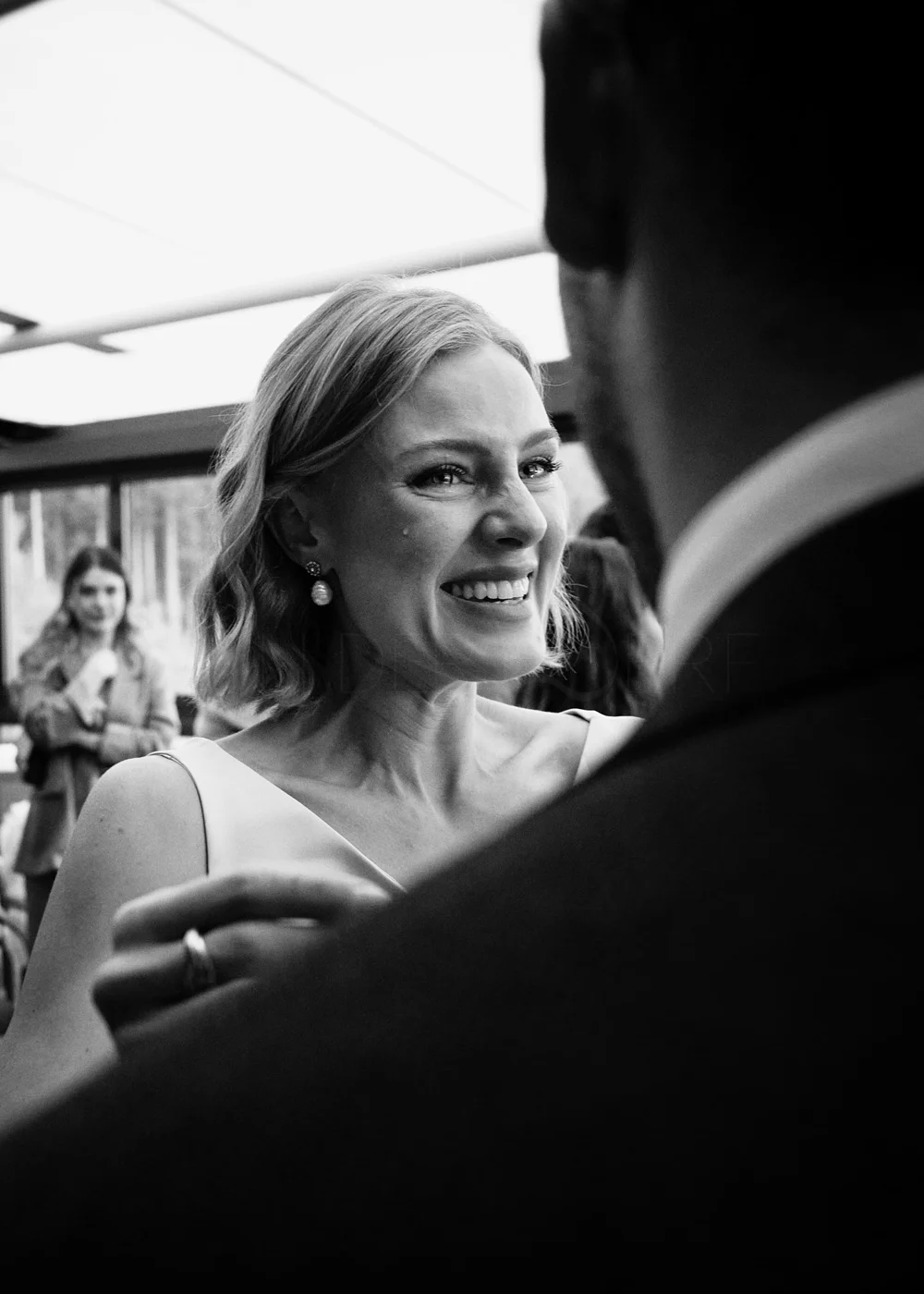 Black and white photo of a smiling woman with short, wavy hair, wearing a sleeveless dress and earrings, talking to a person in a suit at what looks like a social event.