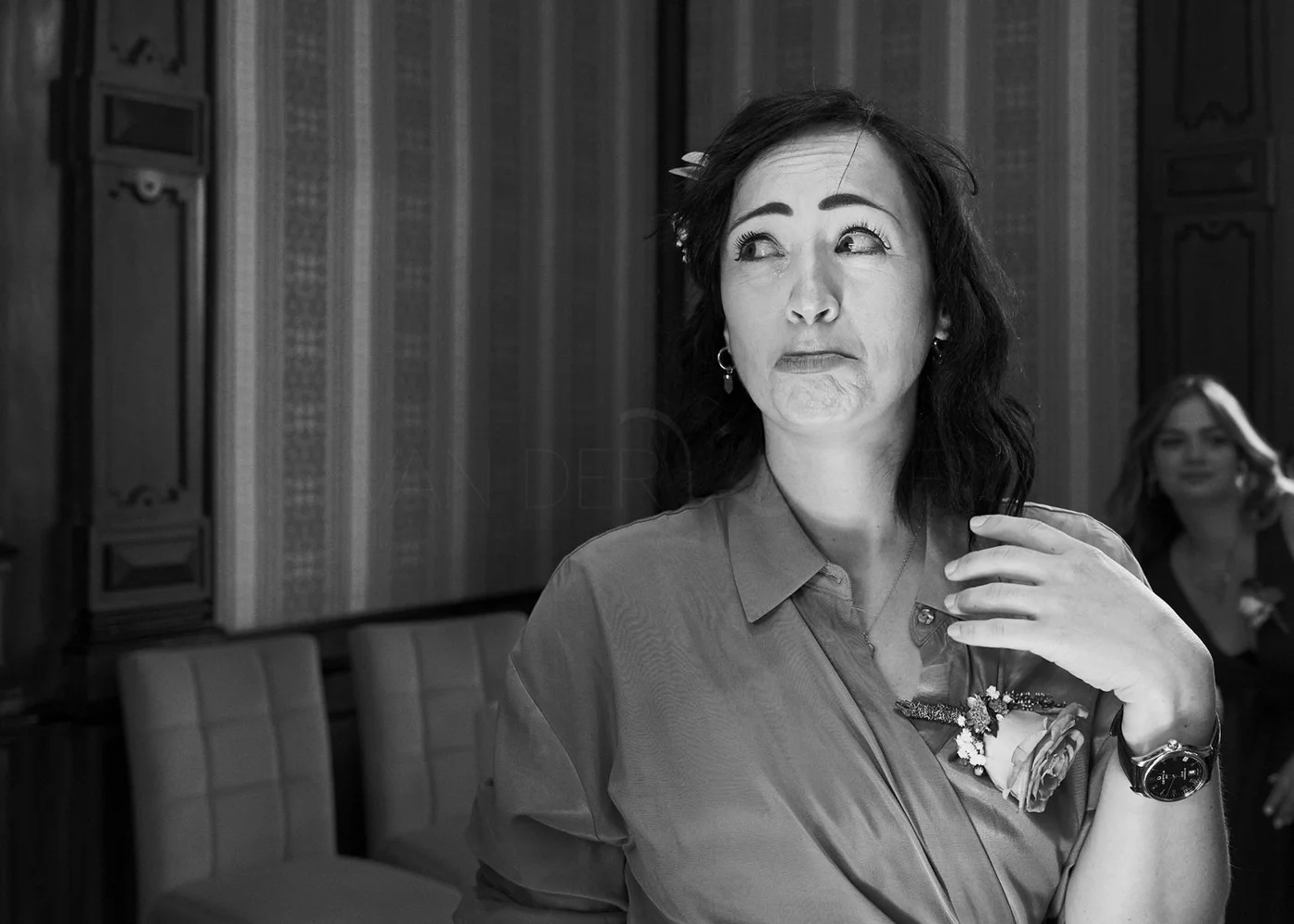 Black and white photo of a woman with dark hair, wearing a shirt with a flower corsage, looking to her right with a playful, unsure expression, while another woman in the background looks on.