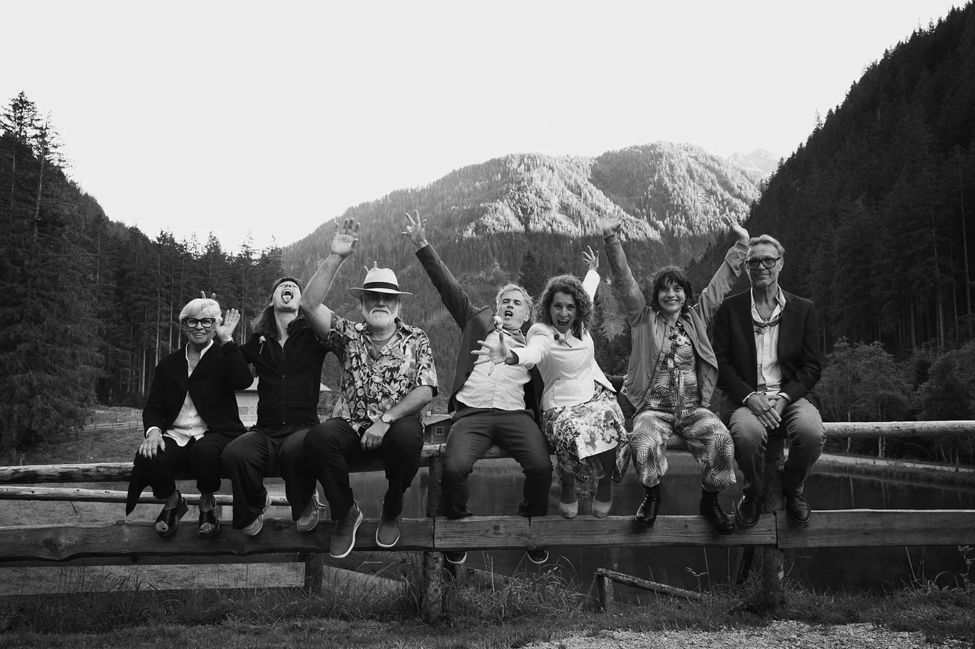 A group of seven people sitting on a wooden railing outdoors, with mountains and dense trees in the background, smiling and raising their hands in a cheerful pose; black and white photo.