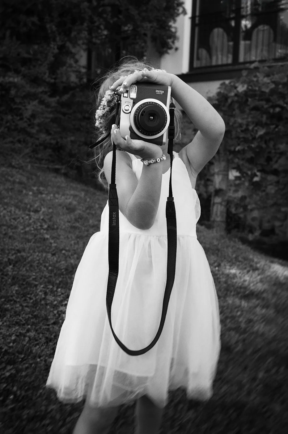 A young girl wearing a dress and a bracelet, holding an instant camera, taking a picture outdoors with trees and a building in the background.