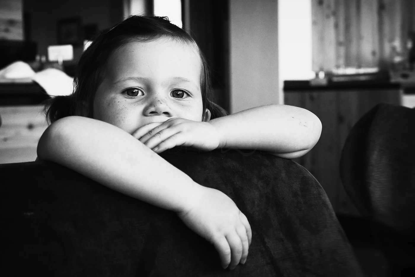 A young girl with pigtails and freckles resting her chin and arm on the back of a couch, looking directly at the camera with a neutral expression in a cozy indoor setting.