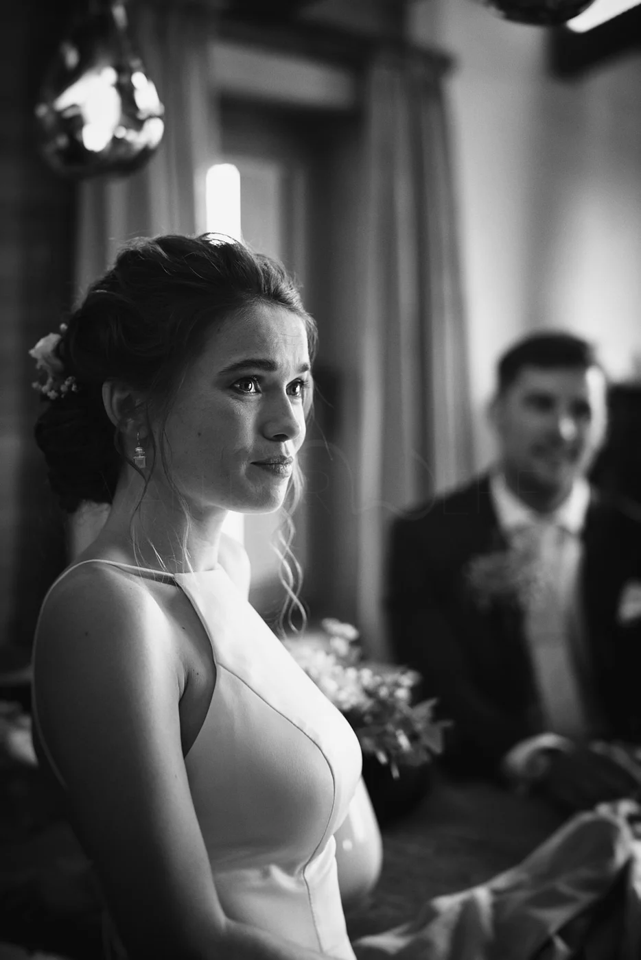 Black and white photo of a woman in a wedding dress with a man in a tuxedo in the background, indoors with curtains and a window.