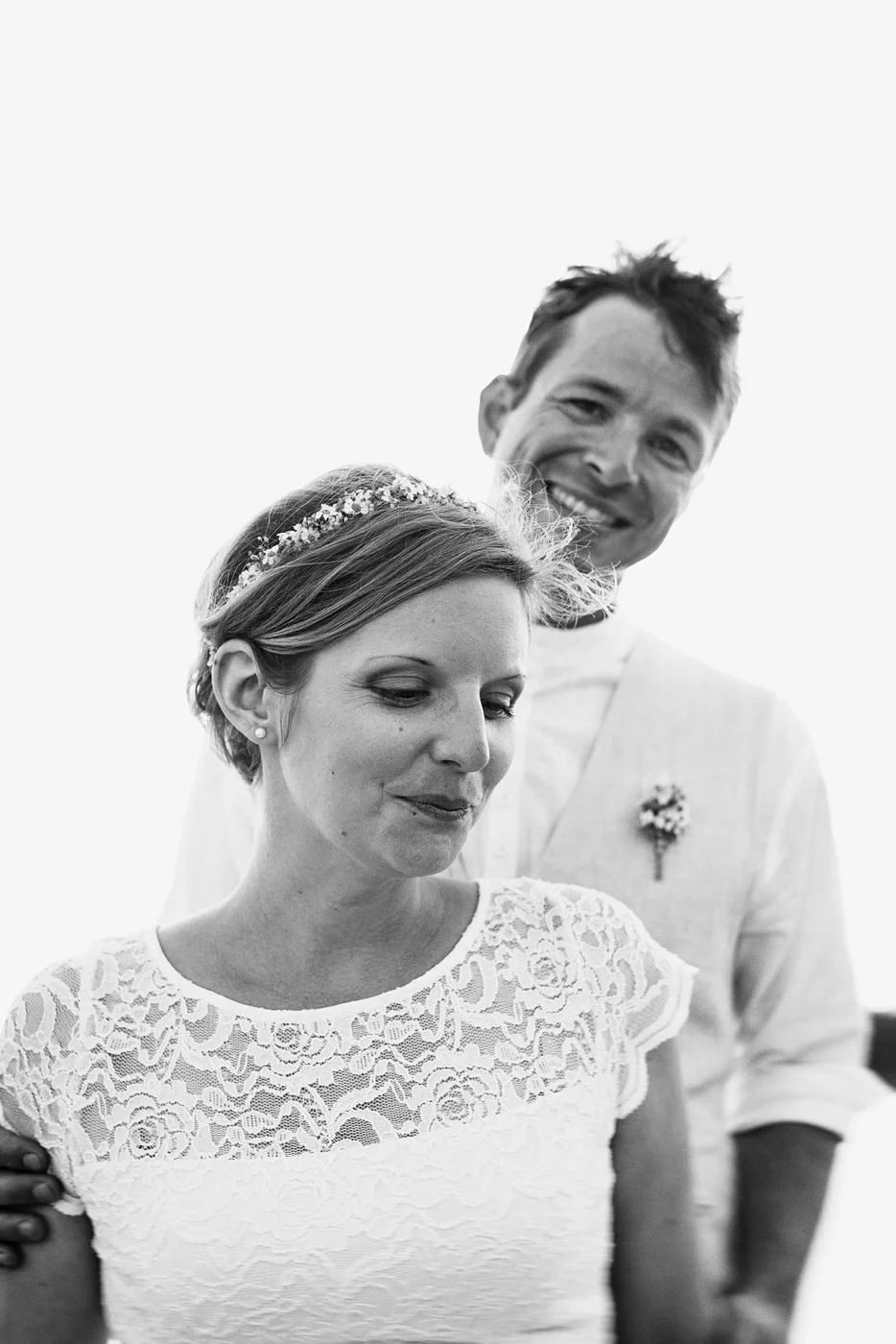 Black and white photo of a woman with short hair wearing a lace dress and a floral headband, with a smiling man with tousled hair standing behind her. Both are dressed in wedding attire.