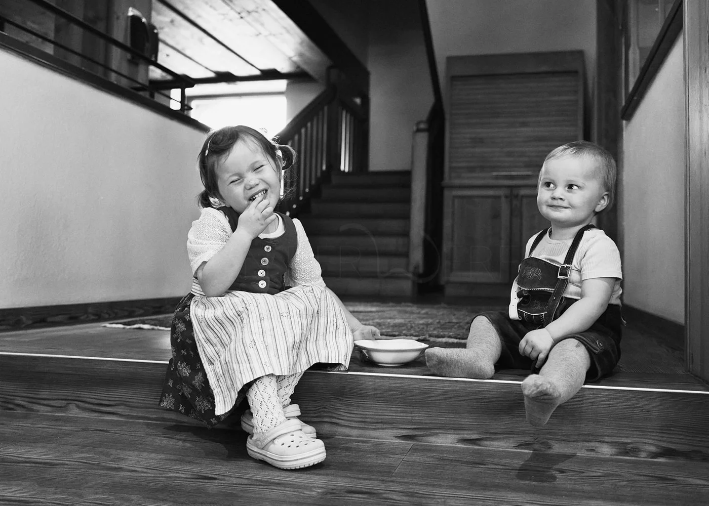 Black and white photo of two young children sitting on a wooden floor in a home; a girl on the left laughs and eats, wearing a dress and Crocs, and a boy on the right looks at her with a slight smile, wearing a T-shirt and overalls.